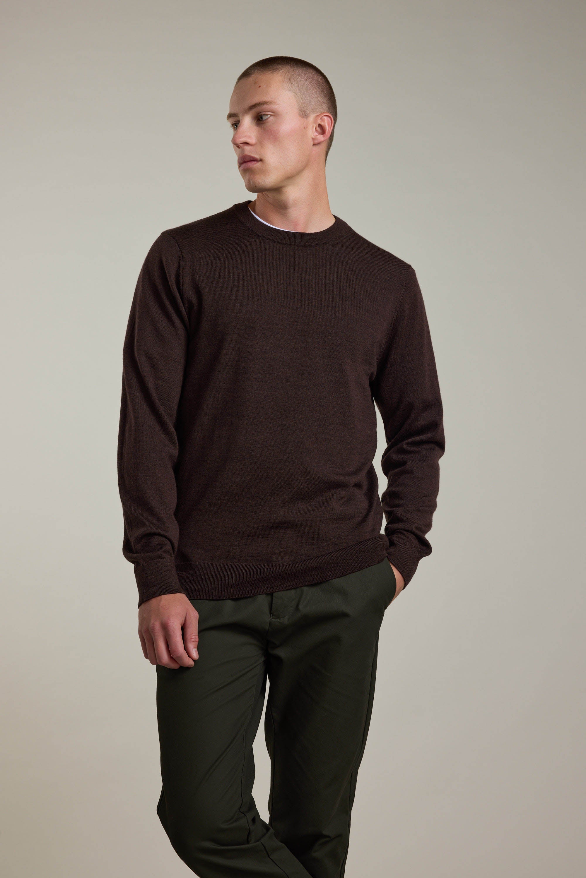 A person stands against a plain background, wearing a dark brown Barkers Skyland ZQrx Merino Crew made from sustainable New Zealand merino wool and dark green pants, with one hand in their pocket and looking to the side.