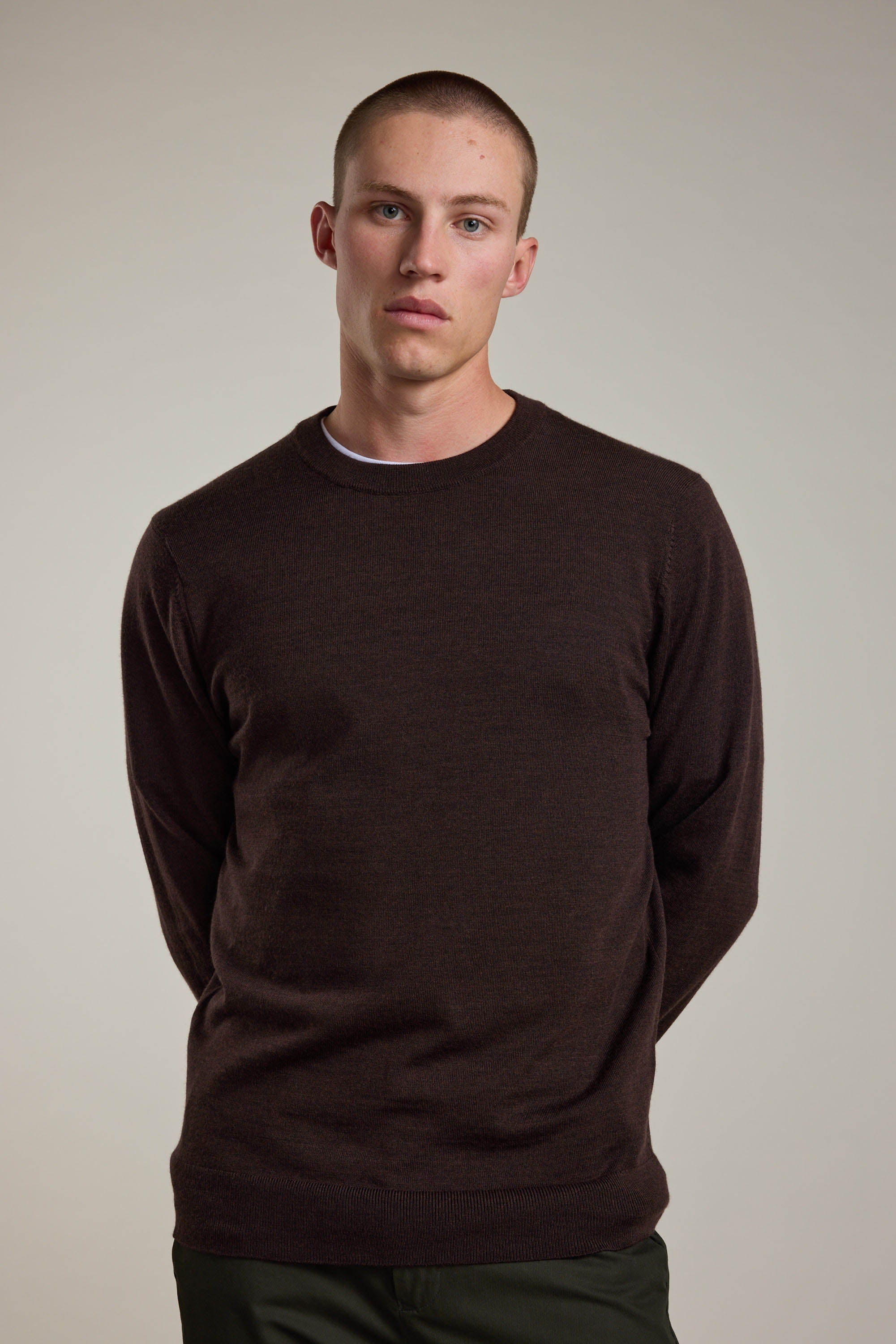 A young man with short hair wears a dark brown Skyland ZQrx Merino Crew by Barkers, made from sustainable merino wool, standing against a plain light background with a neutral expression and his hands behind his back.