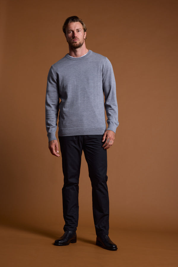 A man stands against a plain brown background wearing the Barkers Skyland ZQrx Merino Crew, a light gray sweater made from ethically sourced merino. He showcases sustainable style with a relaxed posture and neutral expression.