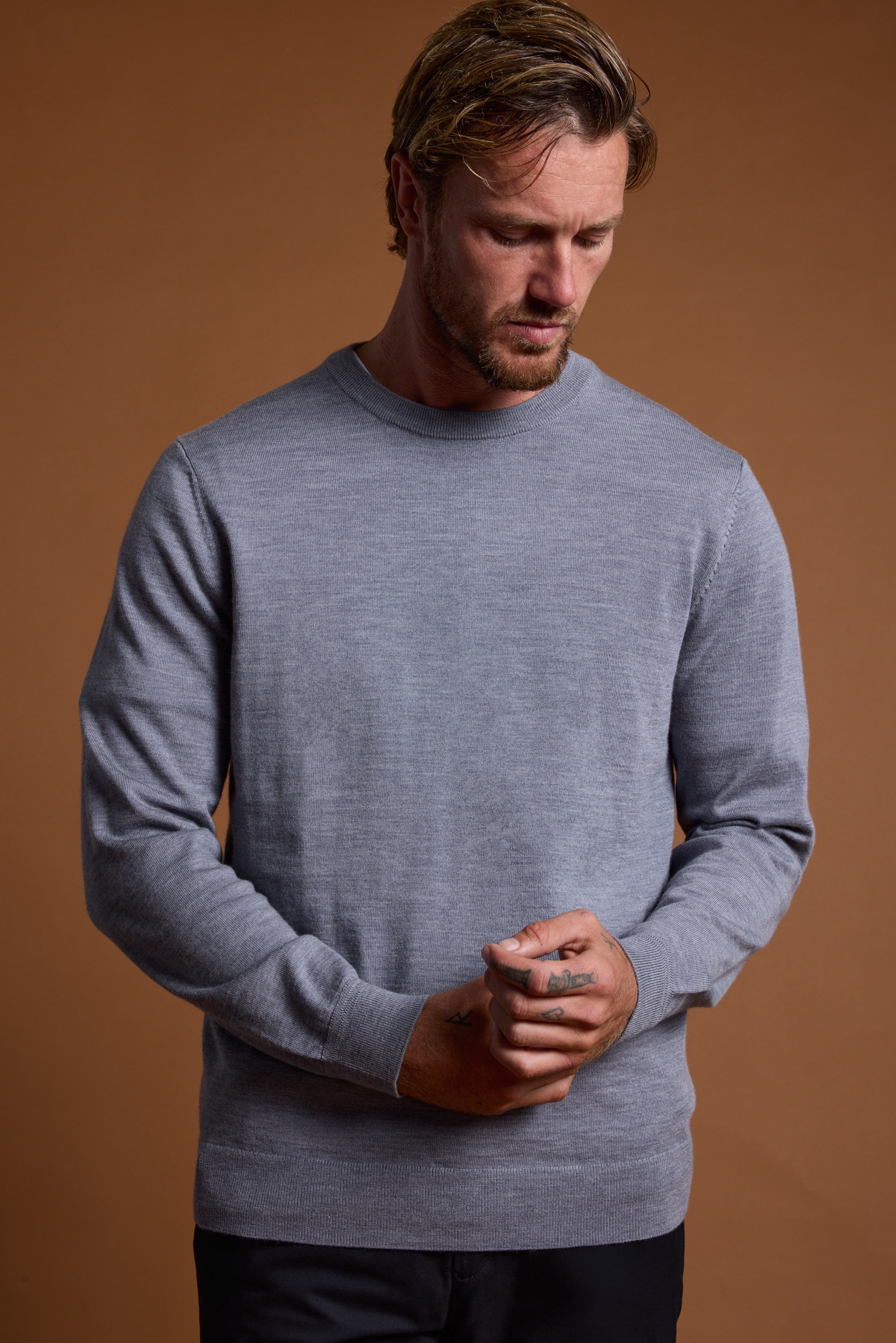 A man with light brown hair and beard wears the Barkers Skyland ZQrx Merino Crew sweater in light gray, standing against a brown background, looking down with hands clasped in front.