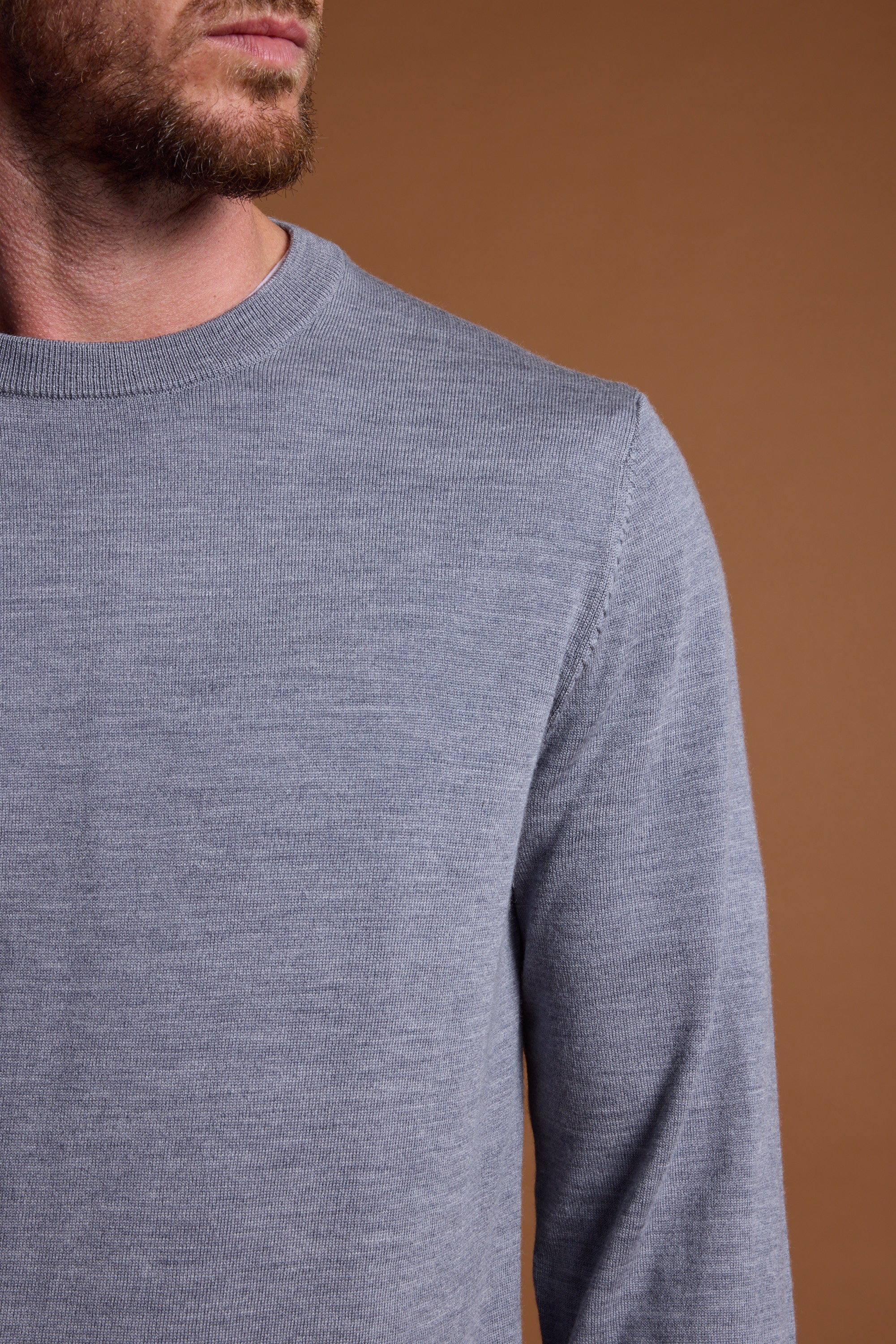 A man models the Barkers Skyland ZQrx Merino Crew, a light gray sweater made from ethically sourced merino wool. He stands against a plain brown background, with only his lower face, neck, and upper torso visible.