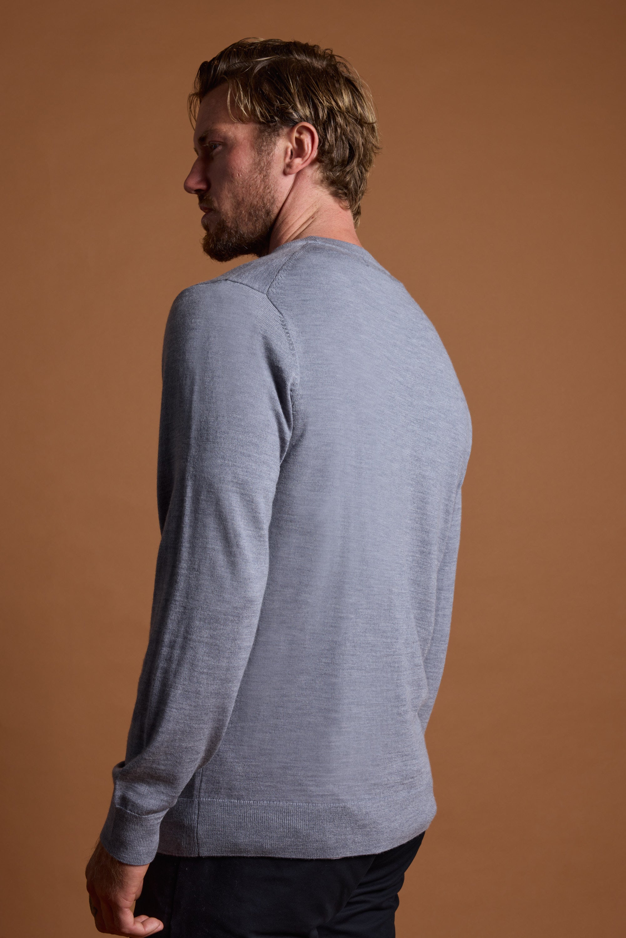 A man with light brown hair and a beard stands sideways, looking left. He is wearing the Barkers Skyland ZQrx Merino Crew, a light gray long-sleeve shirt made from ethically sourced merino, and dark pants against a plain brown background.