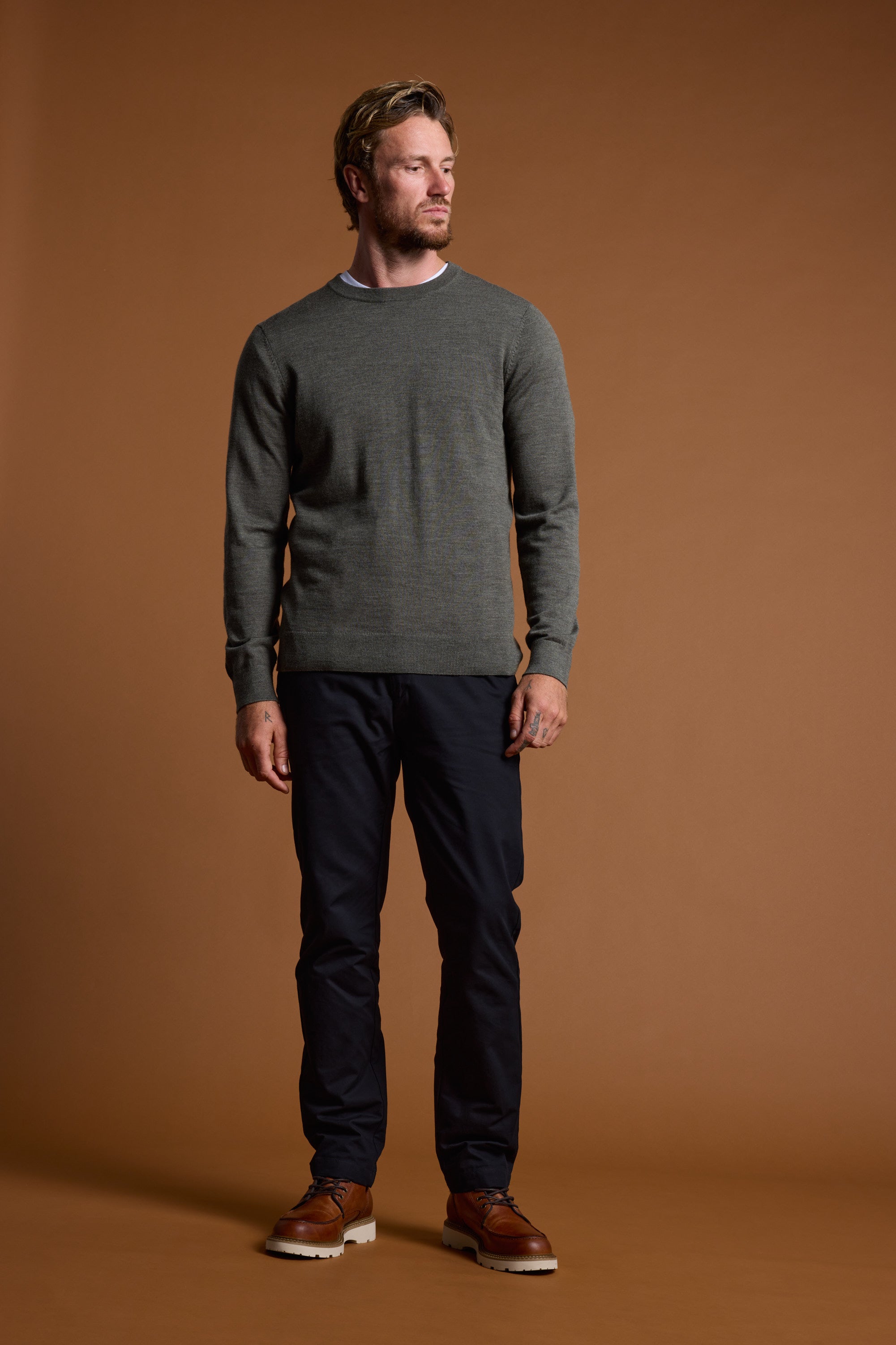 A man with short hair and a beard stands against a plain brown background, wearing the Barkers Skyland ZQrx Merino Crew—a gray sweater made from ethically sourced merino—showcasing a modern, sustainable style.
