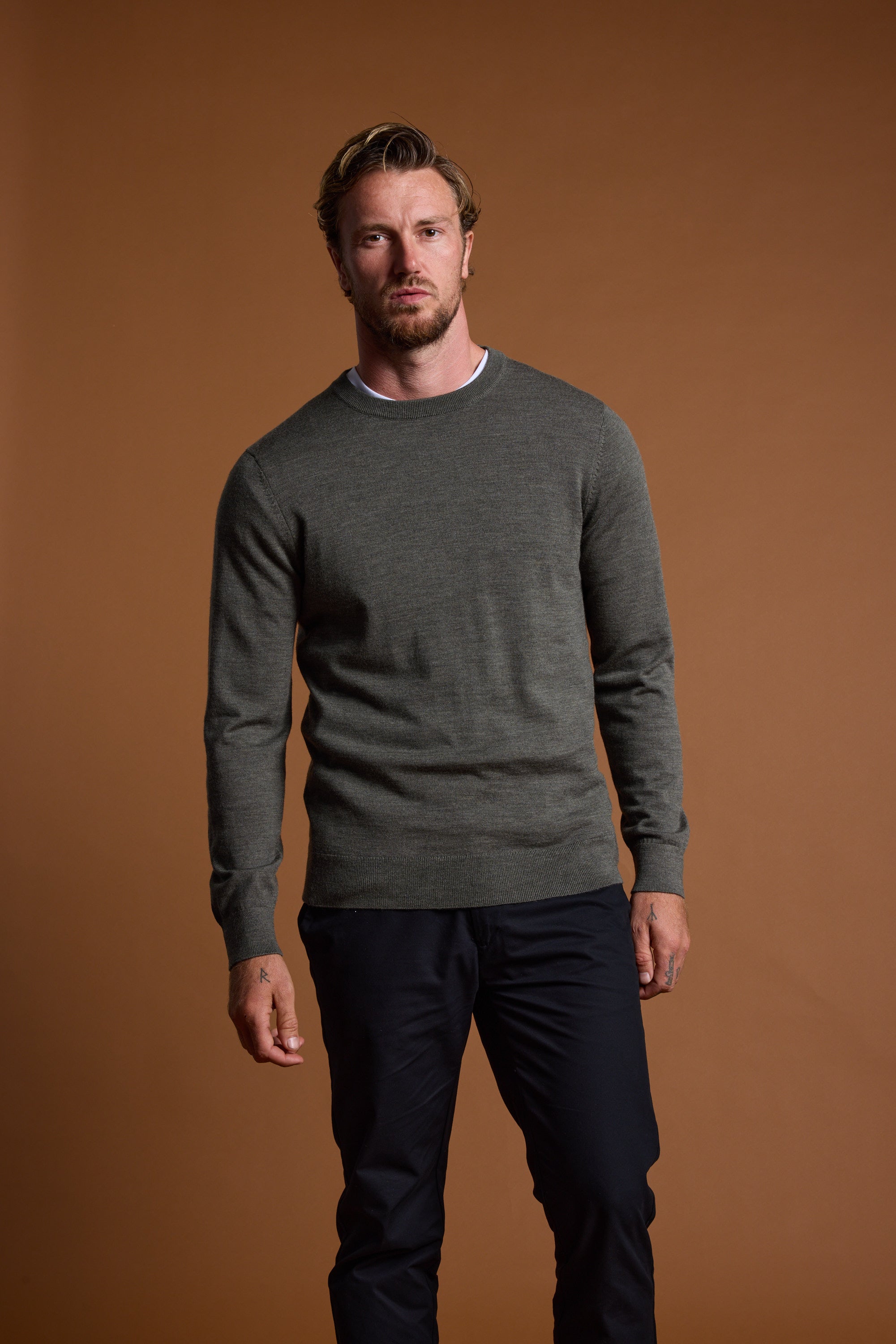 A man with light brown hair and a beard stands against a brown backdrop, wearing the Barkers Skyland ZQrx Merino Crew sweater in gray over a white shirt and dark pants, with a neutral expression and his left hand slightly clenched.
