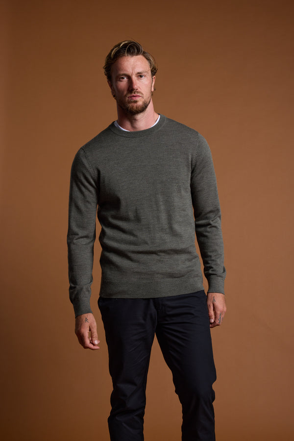 A man with light brown hair and a beard stands against a brown backdrop, wearing the Barkers Skyland ZQrx Merino Crew sweater in gray over a white shirt and dark pants, with a neutral expression and his left hand slightly clenched.
