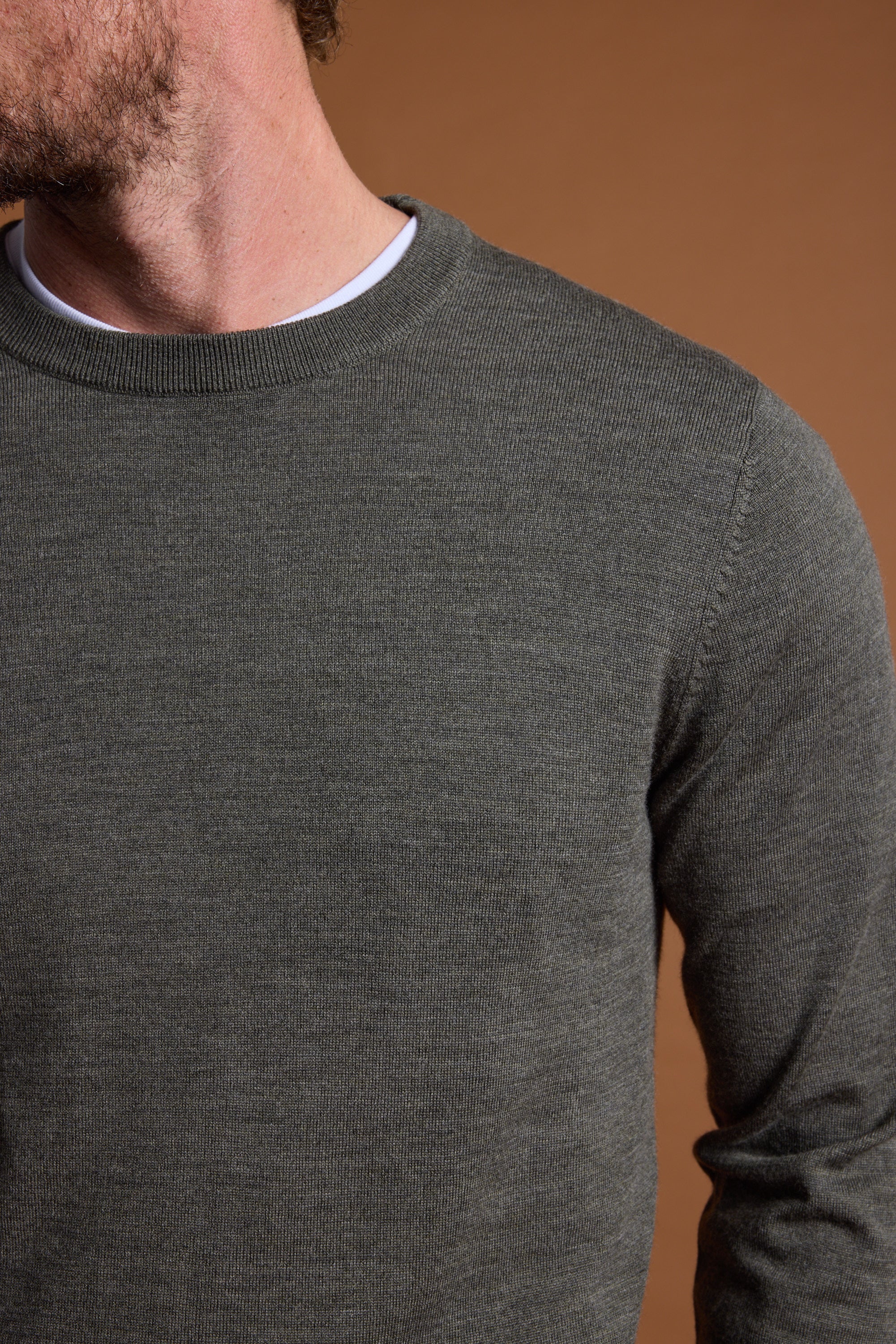 A close-up of a man wearing the Barkers Skyland ZQrx Merino Crew in dark green over a white tee, shown shoulder to mid-torso against a plain brown background, with his face partly out of frame.