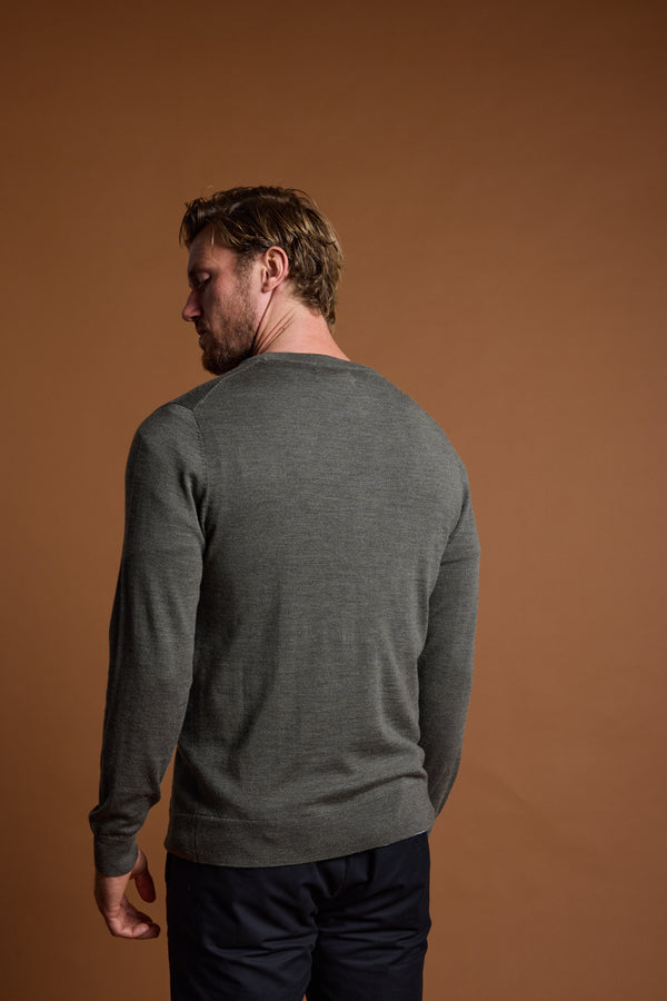 A man with light brown hair, wearing the Barkers Skyland ZQrx Merino Crew in grey and dark pants, stands facing away from the camera against a plain brown background.