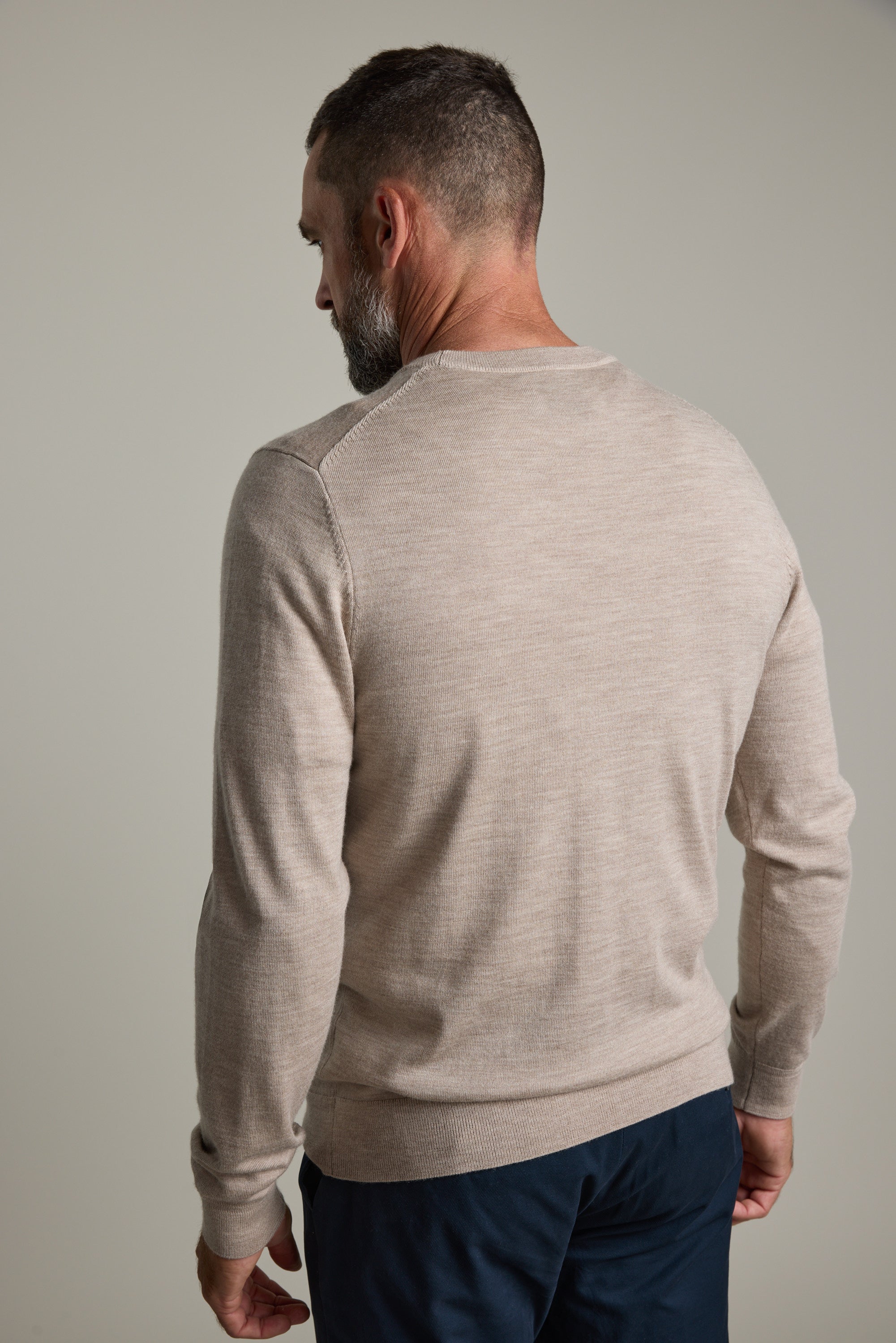 A man in a beige Barkers Skyland ZQrx Merino Crew and dark pants stands against a plain background.