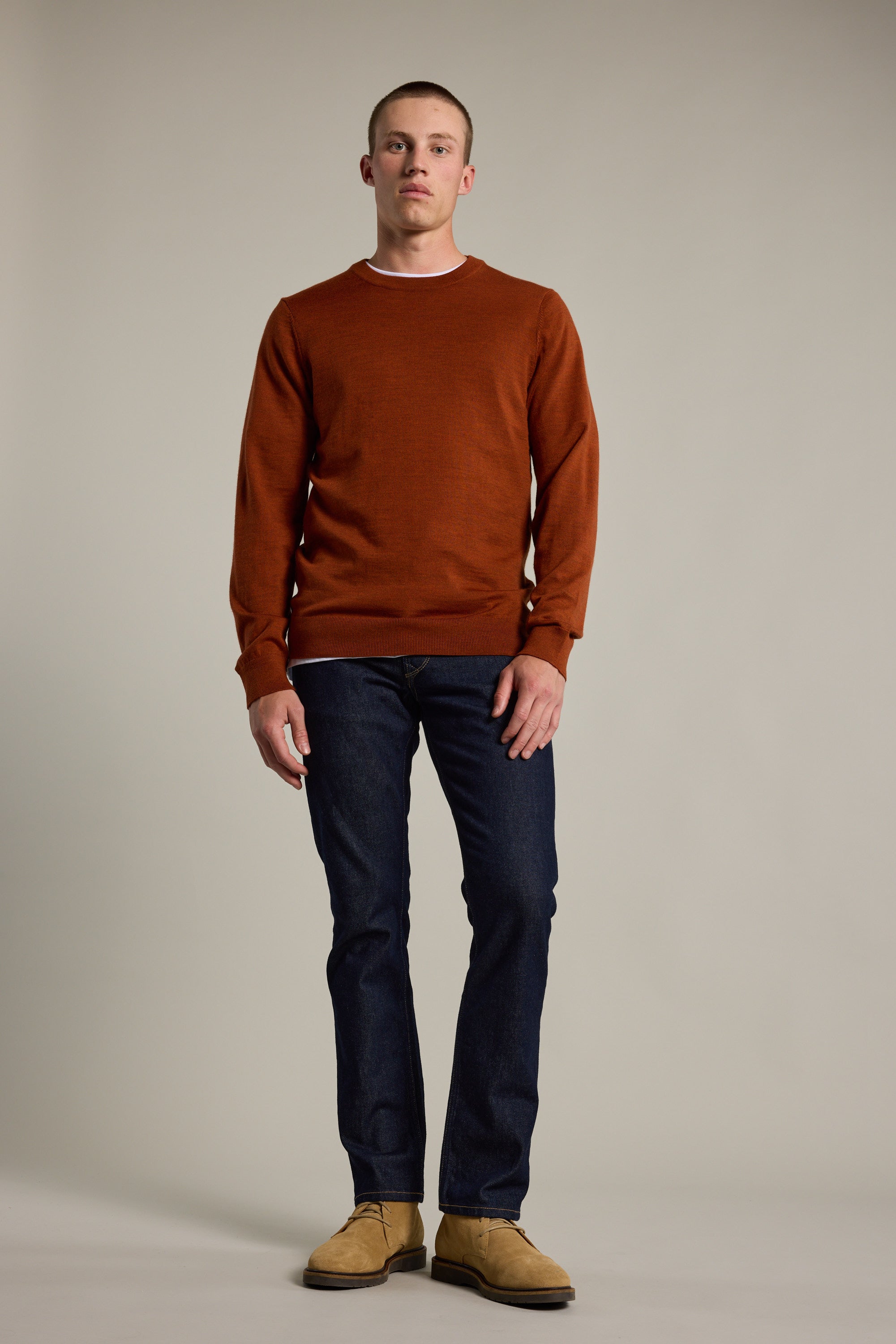 A man faces forward against a plain background, wearing the Barkers Skyland ZQrx Merino Crew in rust, crafted from ethically sourced New Zealand merino wool. He pairs it with dark blue jeans and light brown shoes, arms relaxed by his sides.