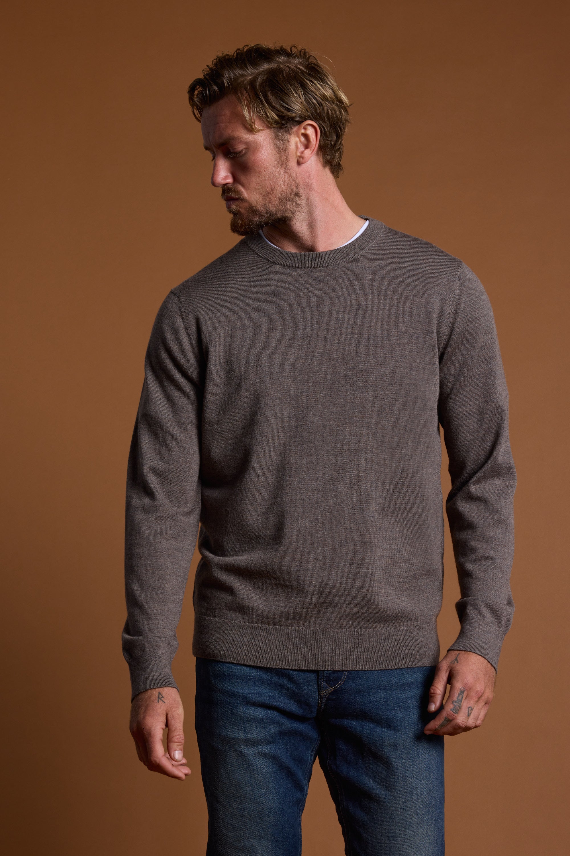 A man with light brown hair and a beard wears the Barkers Skyland ZQrx Merino Crew in brown over a white shirt and blue jeans. He looks down to his left with a neutral expression, standing against a plain brown background.