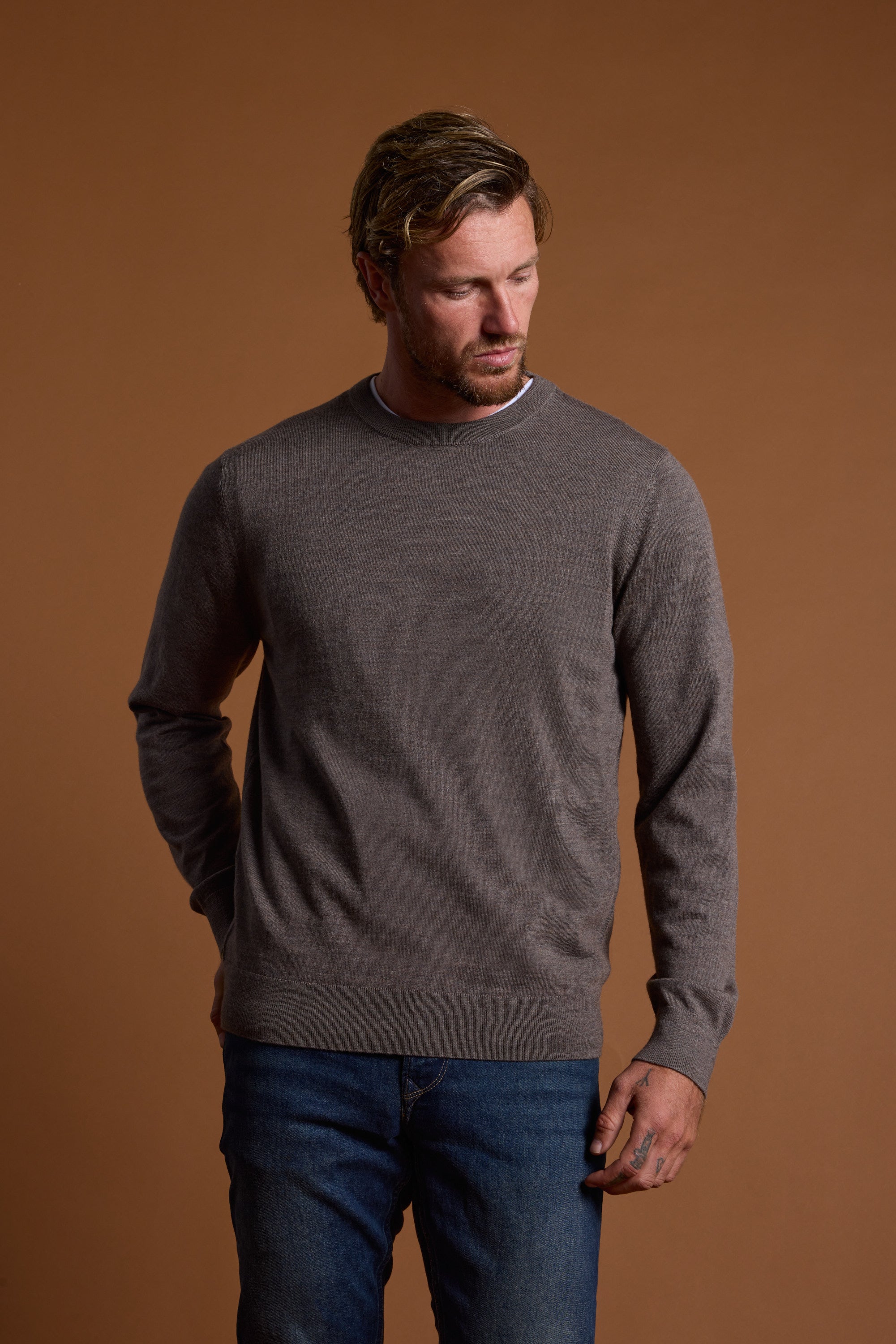 A blond man with a beard stands against a brown background, wearing the Barkers Skyland ZQrx Merino Crew—an ethical, sustainable sweater made from traceable New Zealand merino wool—over a white shirt and blue jeans, looking down to his right.