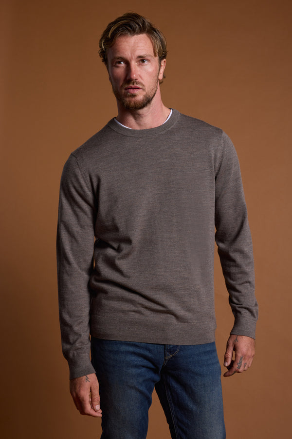 A man with short, wavy hair and facial hair stands against a plain brown background, wearing the Barkers Skyland ZQrx Merino Crew sweater in brownish-gray over a white shirt and blue jeans, looking slightly to the side with a neutral expression.