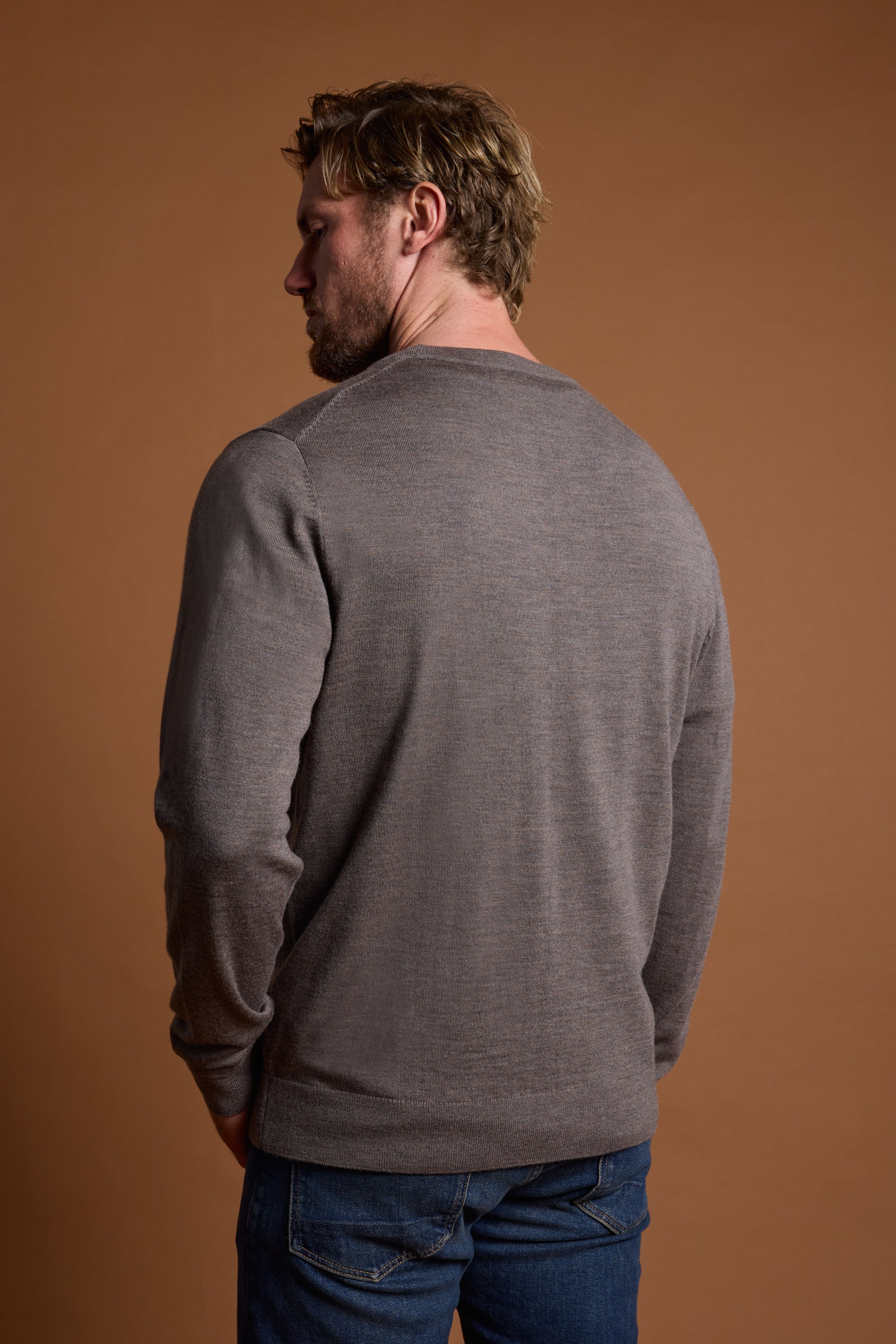 A man with light brown hair stands facing away, wearing the Barkers Skyland ZQrx Merino Crew, made from sustainable traceable wool, paired with blue jeans against a plain brown background.