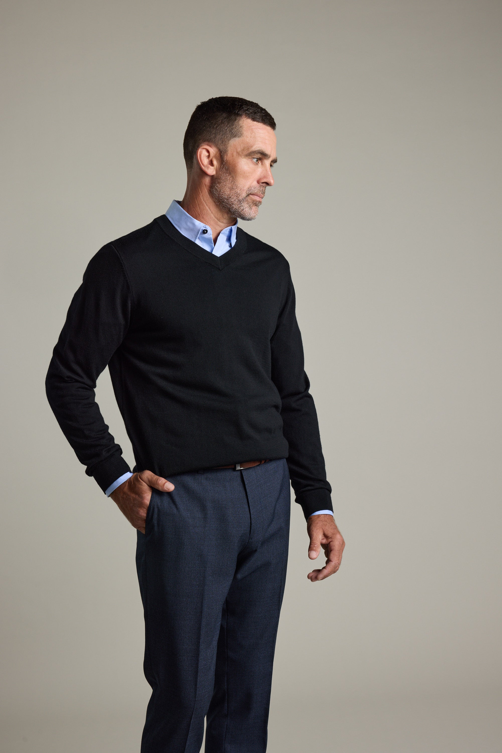 Man in a Barkers Skyland ZQrx Merino Vee sweater over a blue shirt stands by a plain background.