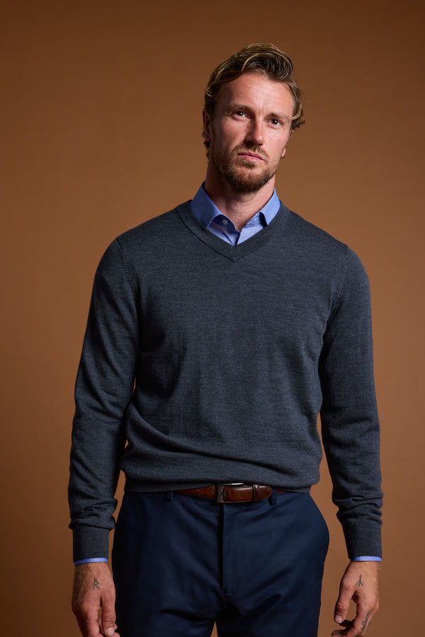 A man with short, wavy blond hair and a beard stands against a brown background, wearing Barkers' Skyland ZQrx Merino Vee sweater over a blue shirt and dark pants, looking seriously at the camera.