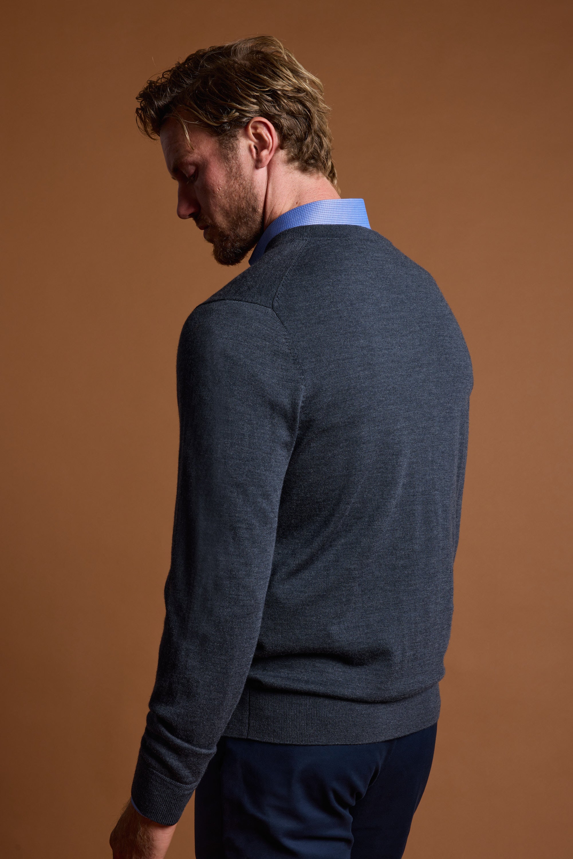 A man with light brown hair, facing away against a plain brown background, wears a blue collared shirt and the Barkers Skyland ZQrx Merino Vee dark gray sweater.