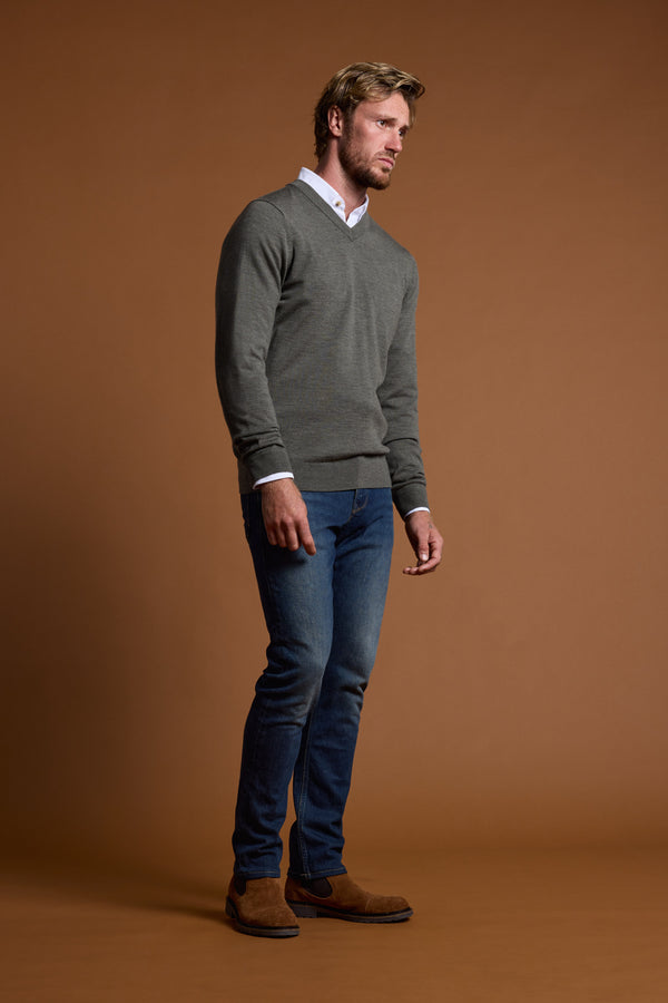A man with light hair and a beard, looking to the side, wears the Barkers Skyland ZQrx Merino Vee sweater over a white shirt, blue jeans, and brown suede shoes against a plain brown background.