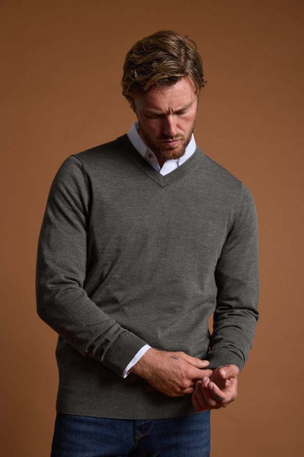 A man with wavy hair wears the Barkers Skyland ZQrx Merino Vee, a grey New Zealand merino wool sweater, over a white collared shirt and blue jeans as he adjusts his sleeve against a plain brown background.