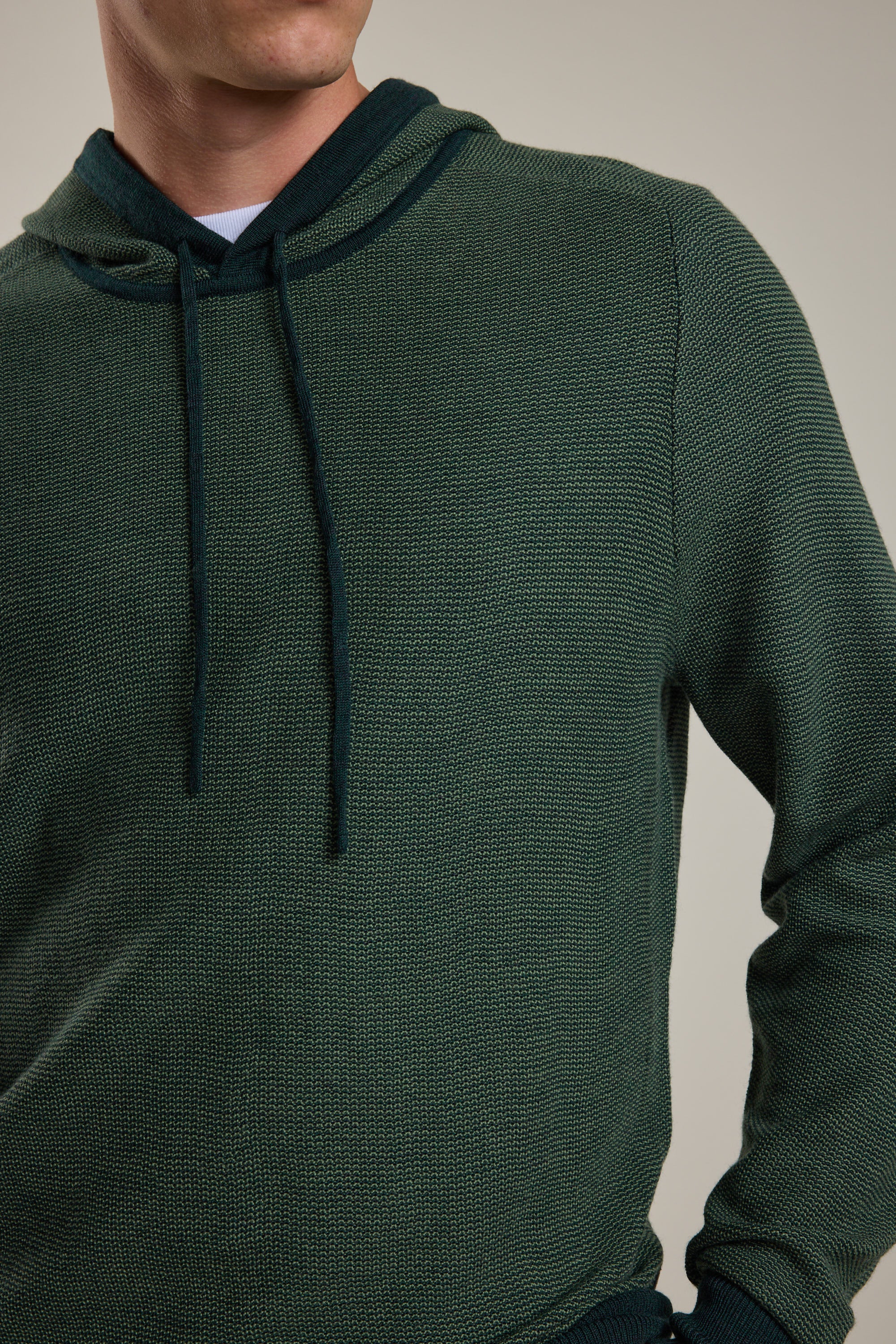 A person wears the Barkers Roxbury Merino Hoody in textured dark green, made from 100% merino wool, shown from shoulders to waist against a neutral background. The face is partially out of frame.