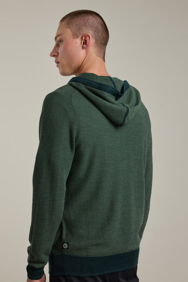A person with short hair wears a dark green Barkers Roxbury Merino Hoody, standing against a plain light background and looking over their left shoulder, showing the back and side profile of the lightweight knit sweater.