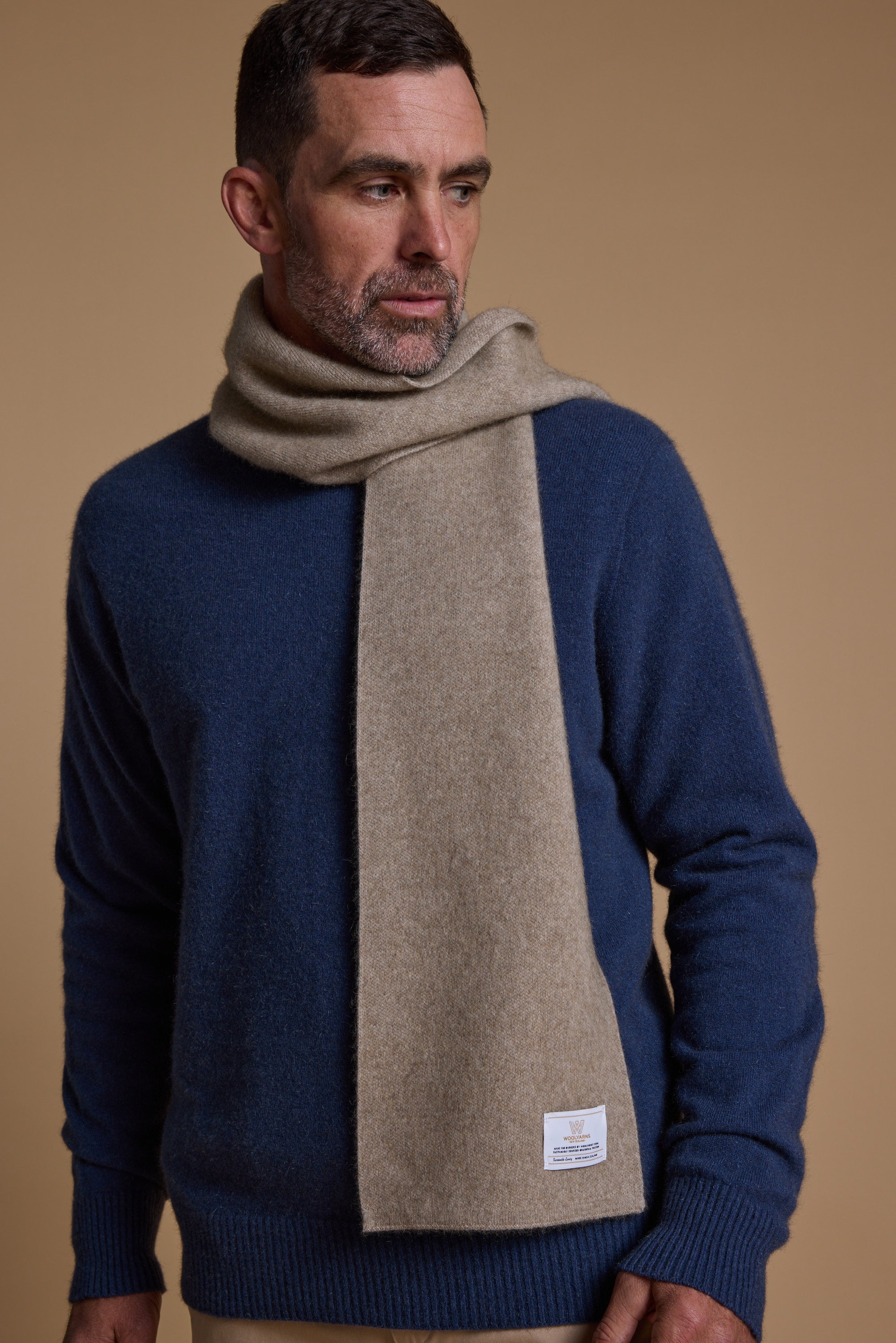 A man with short dark hair and a beard wears a blue sweater and the NZ Made Perino Scarf by Woolyarns Perino, crafted from a merino possum silk blend, against a plain tan background. He looks to his left with one hand at his side.