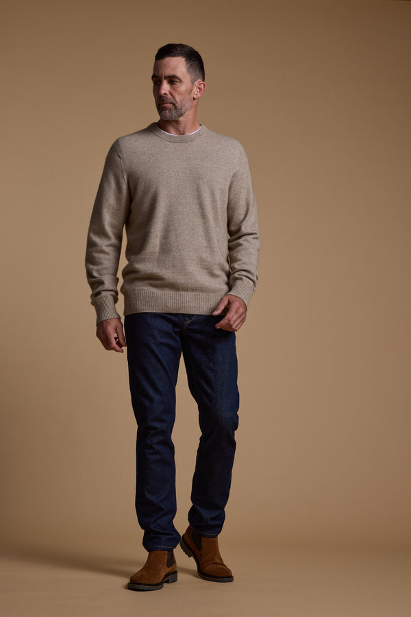 A man with short hair and a trimmed beard stands against a beige background, wearing the Woolyarns Perino NZ Made Perino Crew sweater in light beige, paired with dark blue jeans and brown boots. He looks slightly left with a neutral expression.