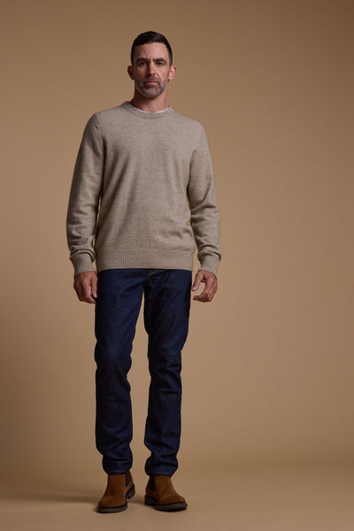 A man with short dark hair and a trimmed beard faces forward against a tan background, wearing the Woolyarns Perino NZ Made Perino Crew, dark blue jeans, and brown boots. He has a neutral expression.