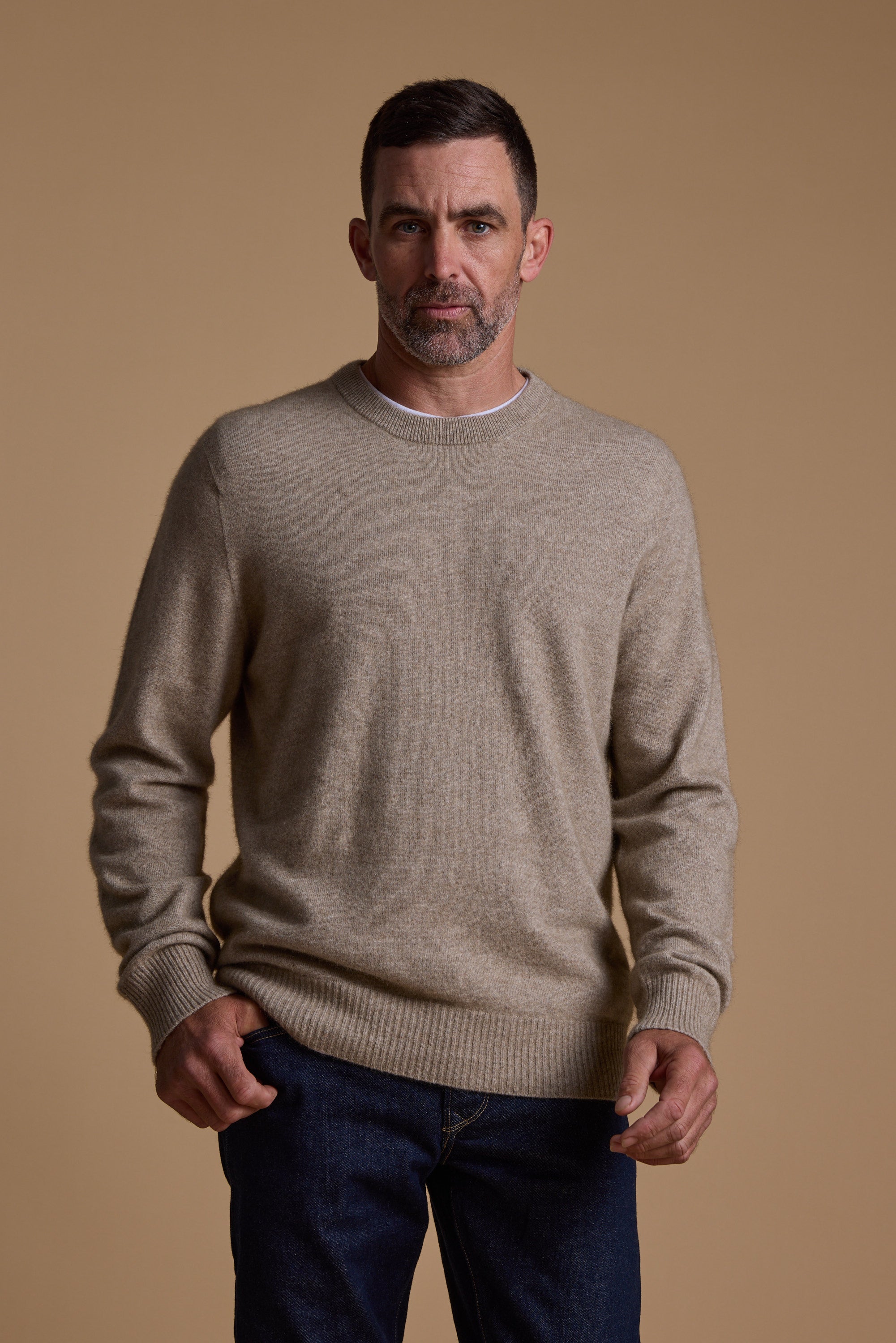 A man with short dark hair and a trimmed beard wears the Woolyarns Perino NZ Made Perino Crew sweater with dark jeans, standing against a plain tan background and looking directly at the camera.