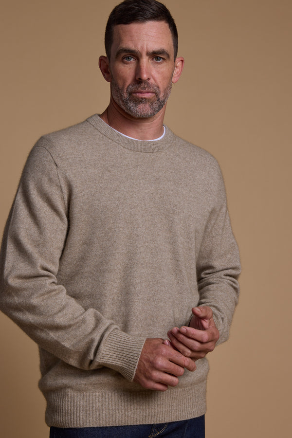 A man with short brown hair and a trimmed beard wears a beige Woolyarns Perino NZ Made Perino Crew over a white shirt, standing against a plain tan background with his hands loosely clasped.
