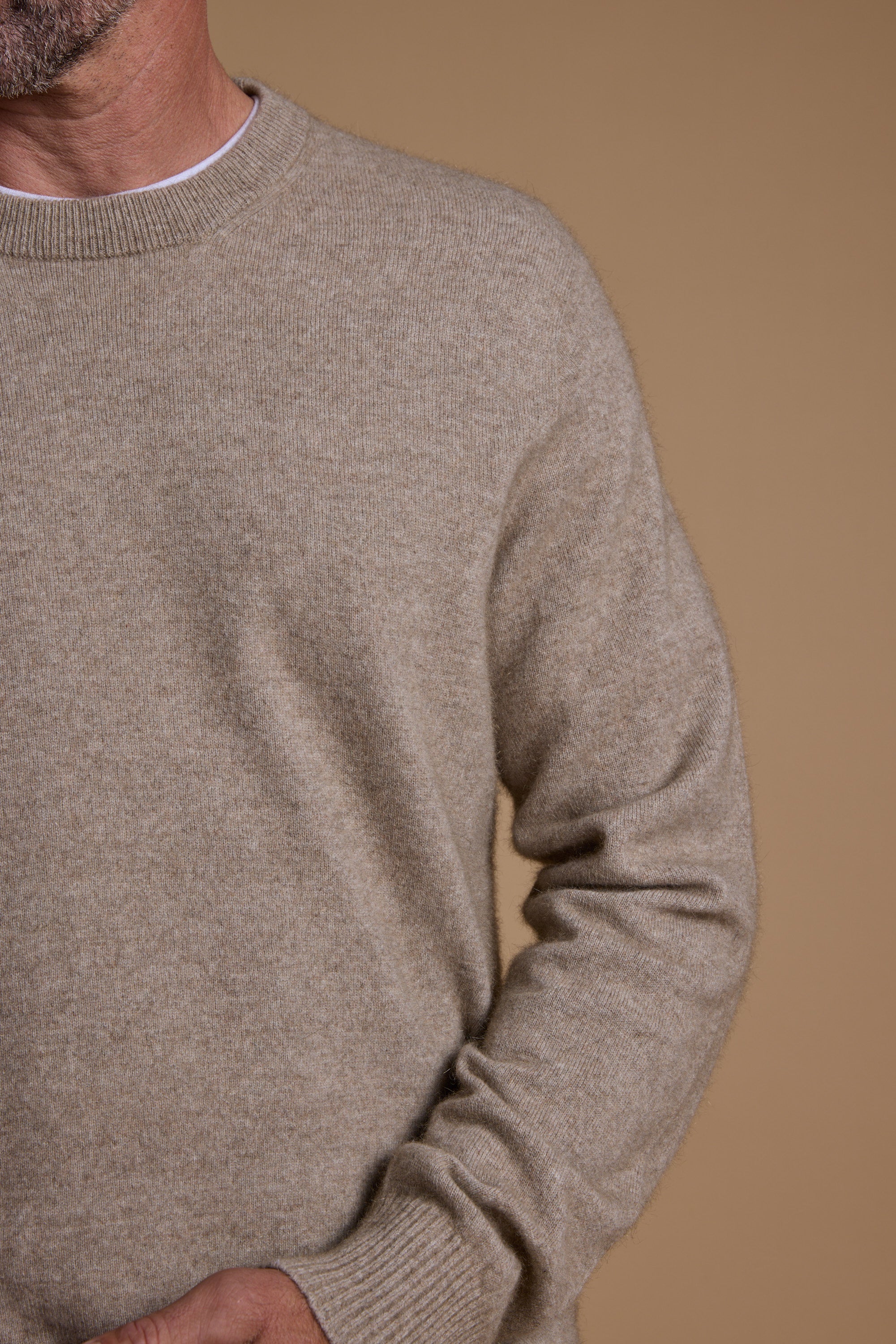 A man models the Woolyarns Perino NZ Made Perino Crew, a beige sweater crafted from luxurious Brushtail Possum yarn and New Zealand merino, worn over a white shirt and shown against a plain tan background.