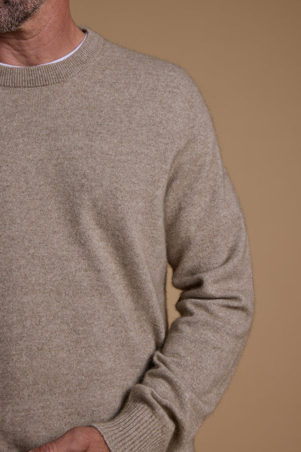 A man models the Woolyarns Perino NZ Made Perino Crew, a beige sweater crafted from luxurious Brushtail Possum yarn and New Zealand merino, worn over a white shirt and shown against a plain tan background.
