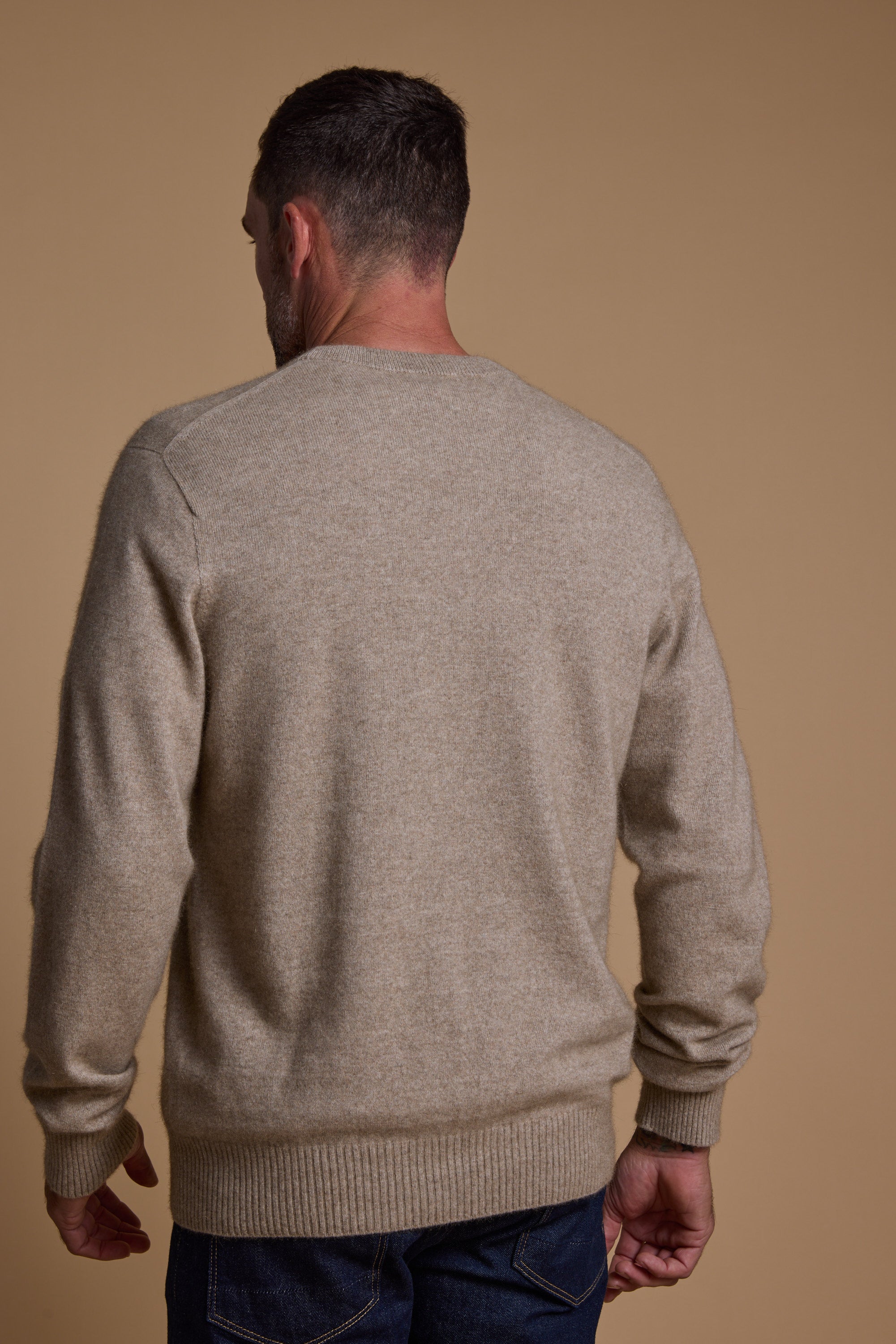 A man with short dark hair stands with his back to the camera, wearing a light beige Woolyarns Perino NZ Made Perino Crew sweater and dark blue jeans, set against a plain tan backdrop.