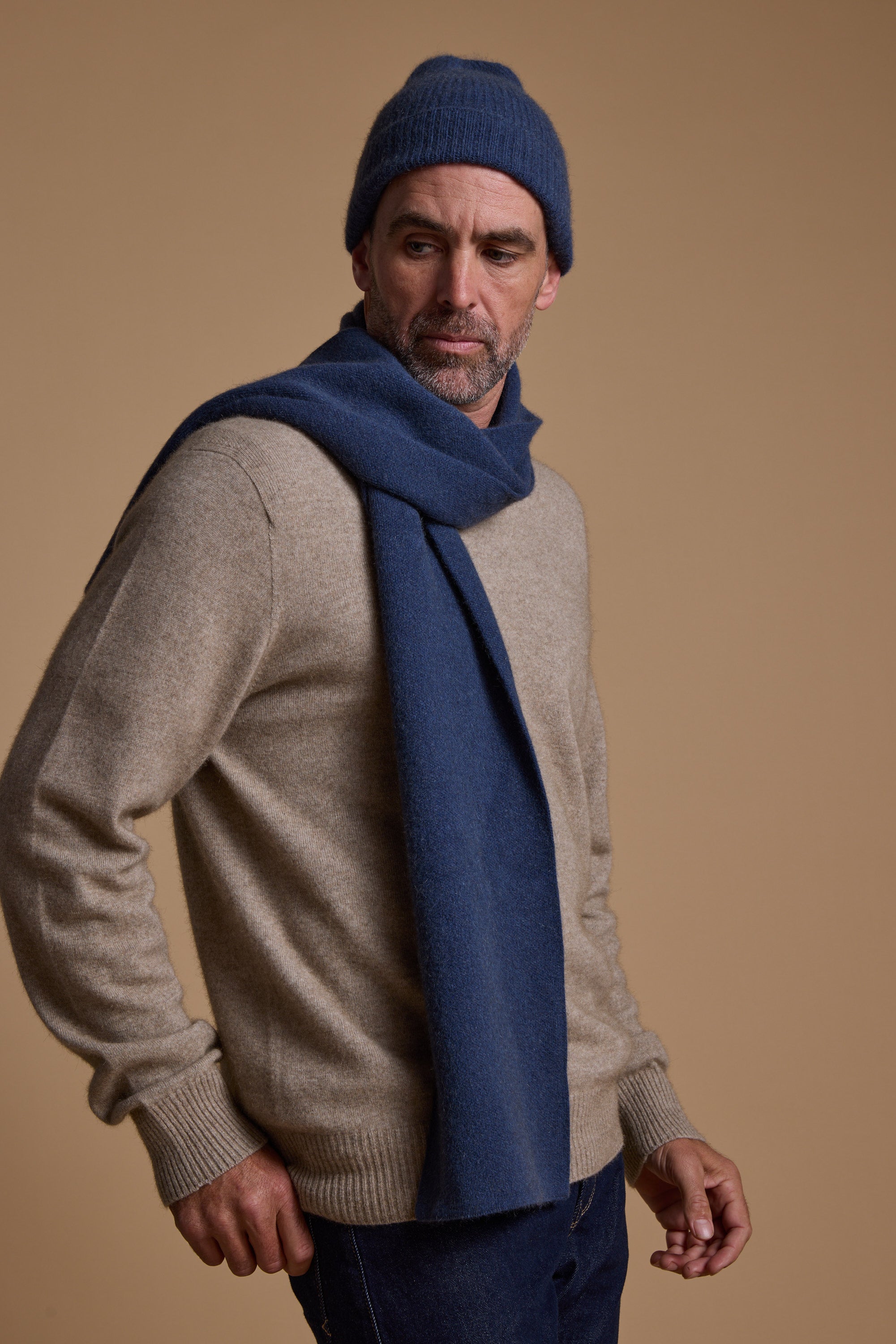 A man in a beige Woolyarns Perino NZ Made Perino Crew sweater, dark blue knit scarf, and matching beanie stands against a neutral background, gazing thoughtfully to the side.