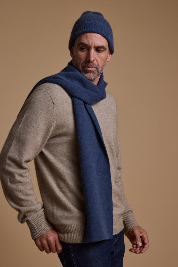 A man in a beige Woolyarns Perino NZ Made Perino Crew sweater, dark blue knit scarf, and matching beanie stands against a neutral background, gazing thoughtfully to the side.