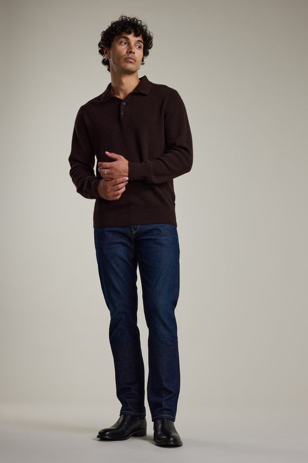 A man stands against a plain background wearing the Barkers Braxton Merino Long Sleeve Polo in dark brown, paired with dark blue jeans and black boots, his hands gently clasped in front of him.
