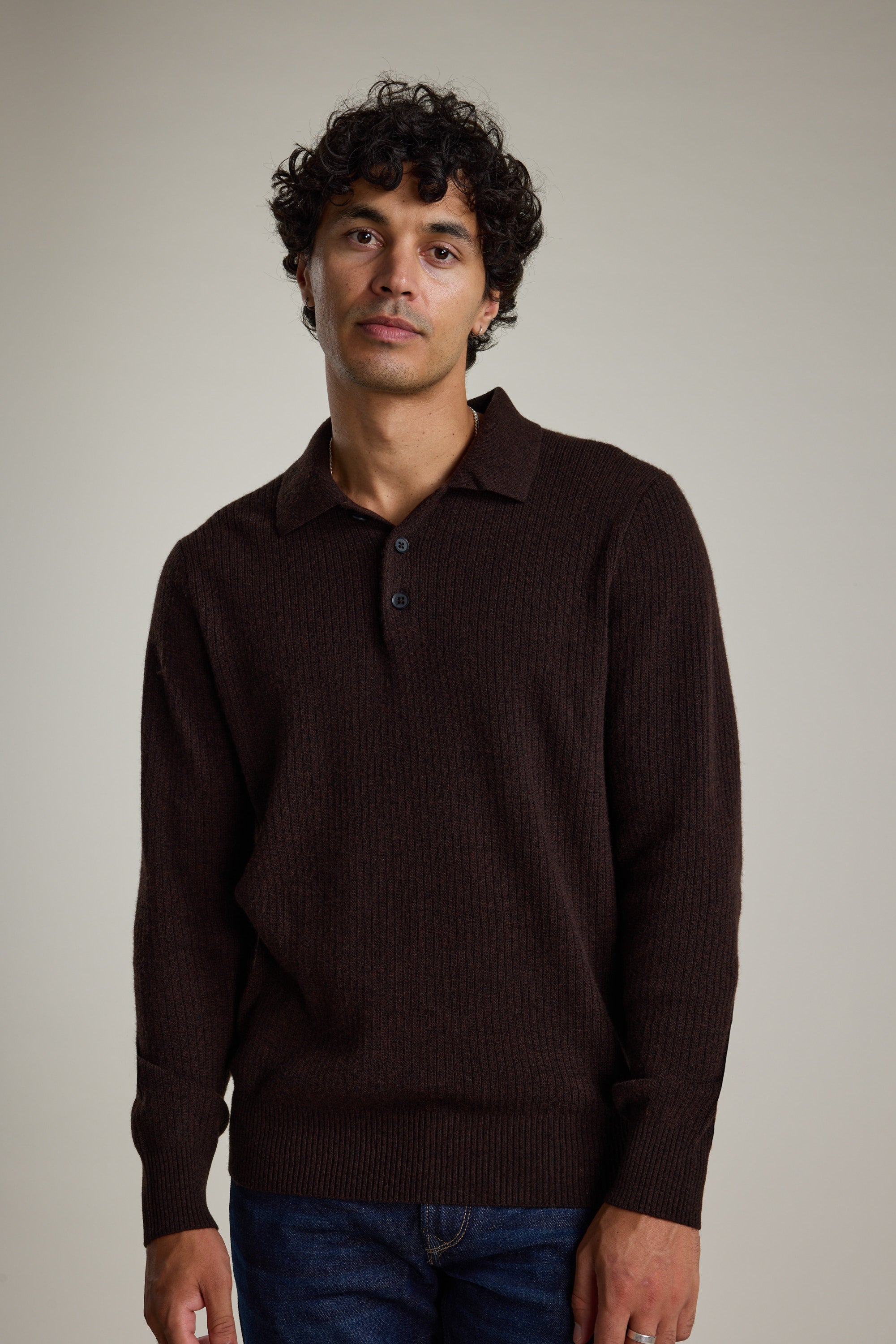 A man with curly dark hair wears the Barkers Braxton Merino Long Sleeve Polo, featuring ribbed detailing and buttons, standing against a plain light background.