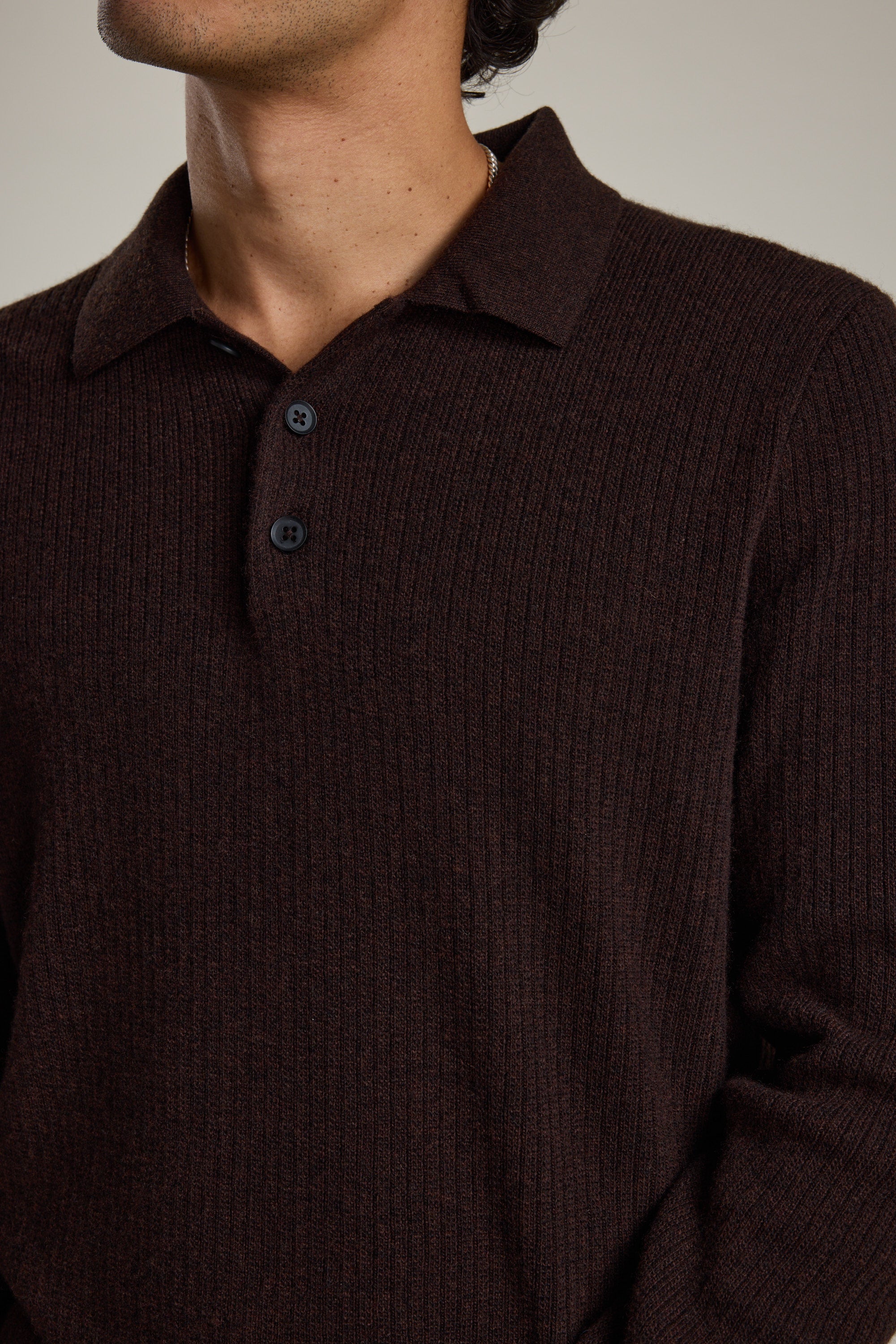 A person wears the Barkers Braxton Merino Long Sleeve Polo in dark brown, featuring ribbed detailing and a classic collar, with two buttons fastened. The image highlights the upper torso and part of the face, emphasizing the polo's texture.