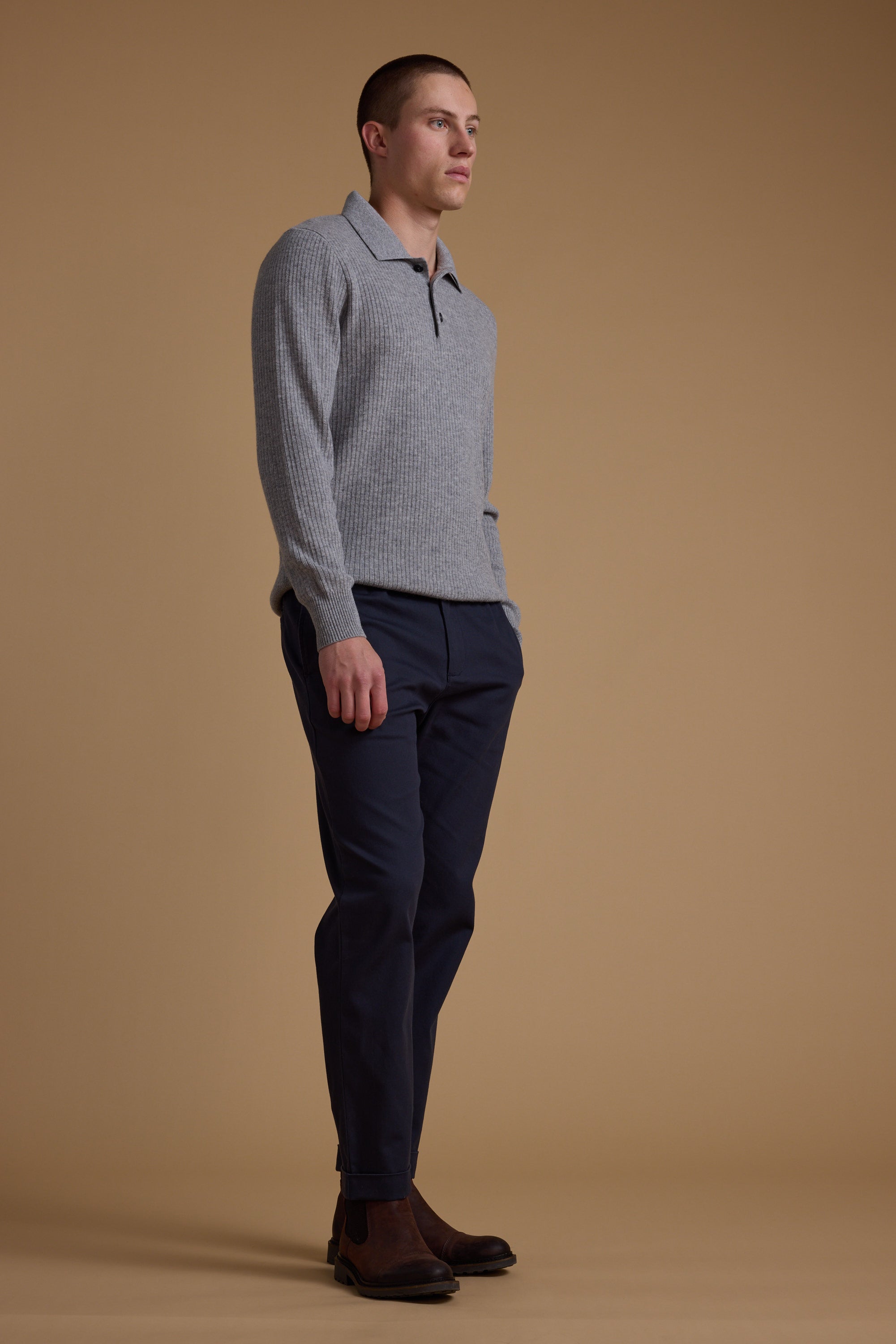 A man stands against a beige background wearing the Barkers Braxton Merino Long Sleeve Polo in light gray, paired with dark navy pants and brown boots. He has short hair, hands in his pockets, and looks slightly to the side.