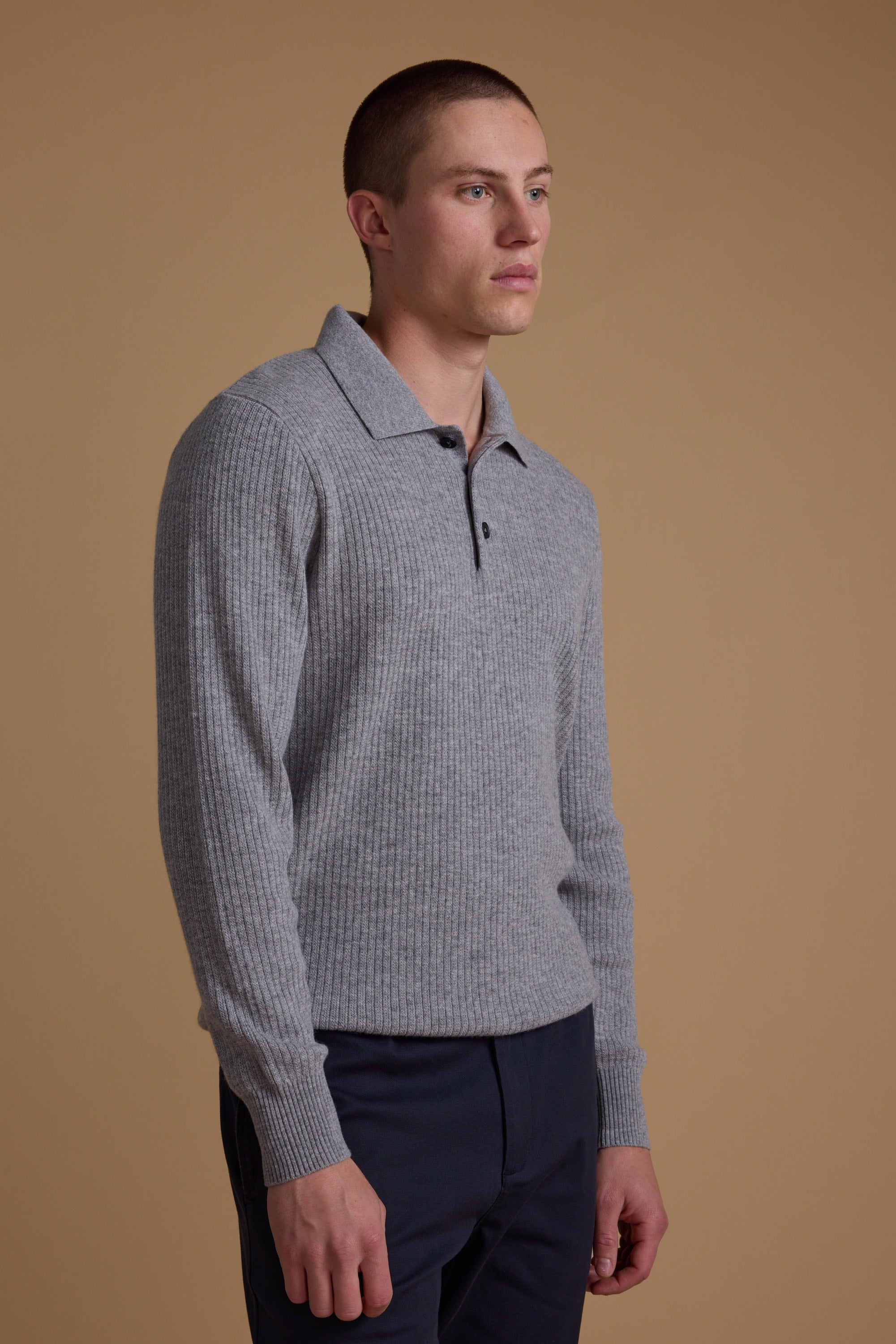 A man with short hair wears the Barkers Braxton Merino Long Sleeve Polo in light grey, featuring ribbed details and buttons, paired with navy blue pants, standing against a plain beige background.