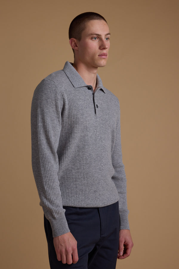 A man with short hair wears the Barkers Braxton Merino Long Sleeve Polo in light grey, featuring ribbed details and buttons, paired with navy blue pants, standing against a plain beige background.