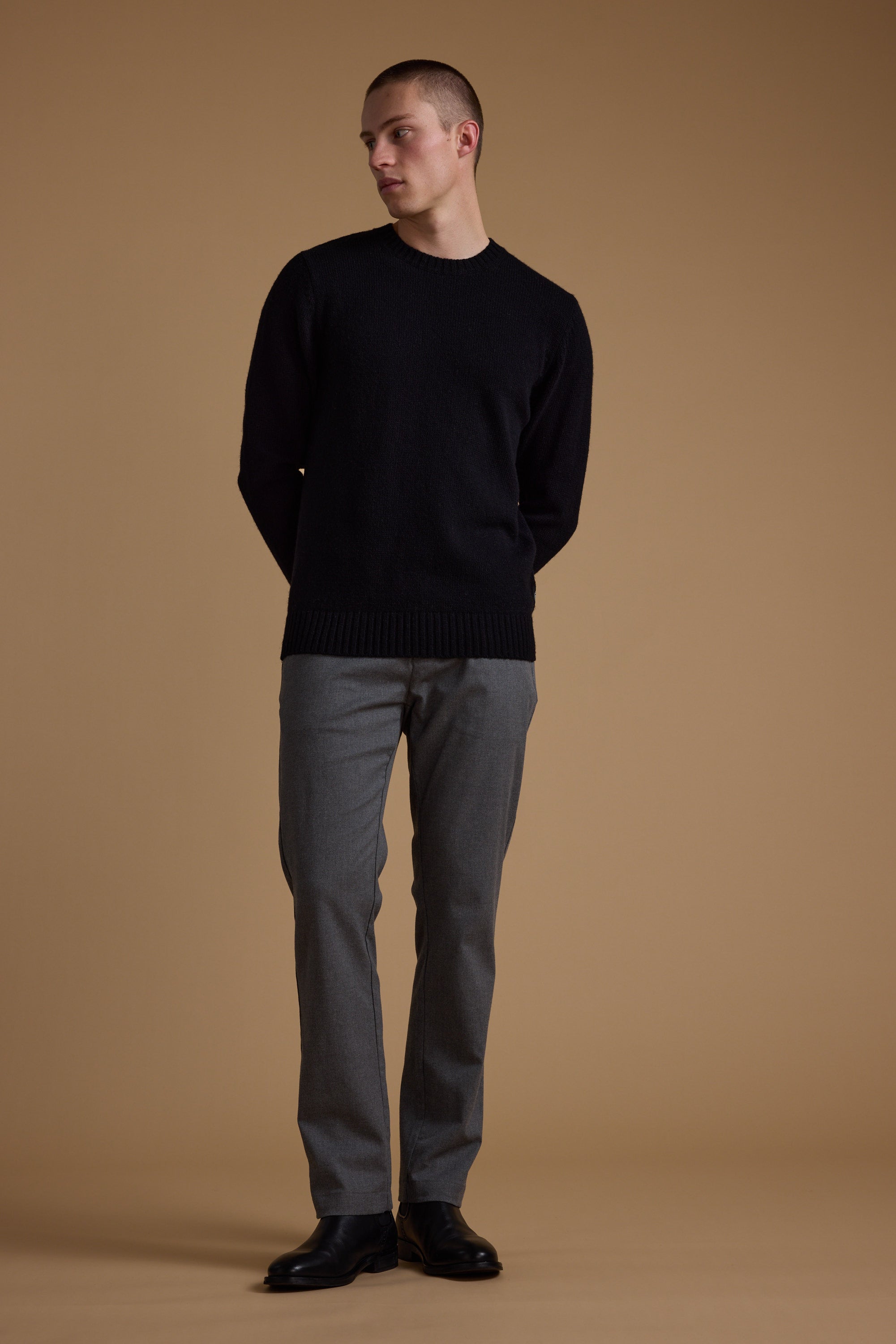 A person stands against a beige background, wearing the Barkers Anglers Merino Crew sweater in black with gray pants and black shoes, hands behind their back and head turned slightly left.