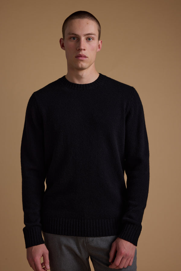 A young man with short hair wears the Barkers Anglers Merino Crew sweater and gray pants, standing against a beige background and looking at the camera—an effortless winter staple.