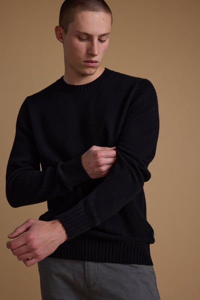 A young man with short hair wears the Barkers Anglers Merino Crew in black and gray pants, standing against a plain beige background, adjusting his left sleeve while looking down.