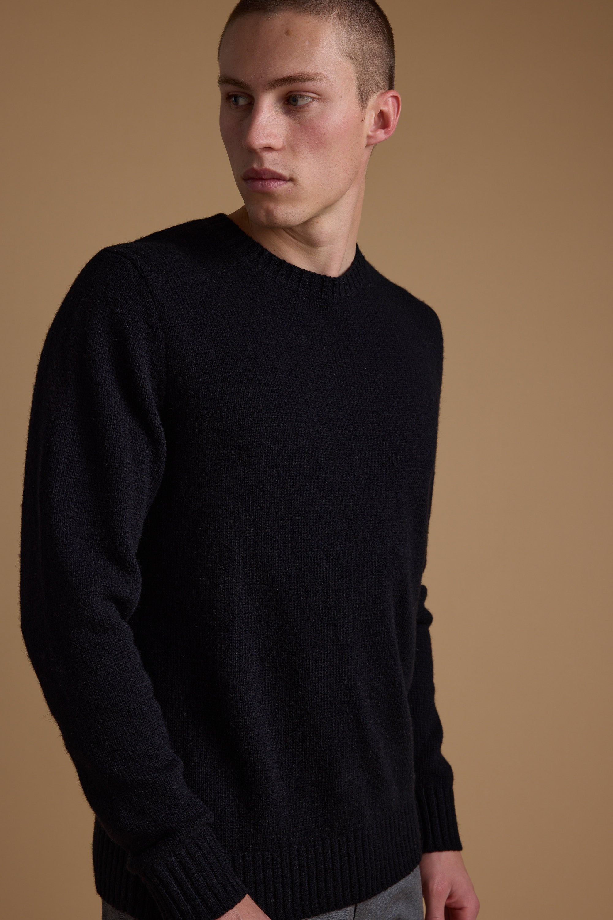 A young man with short hair wears the Barkers Anglers Merino Crew, a classic black merino wool sweater and winter staple, as he looks left against a plain beige background.