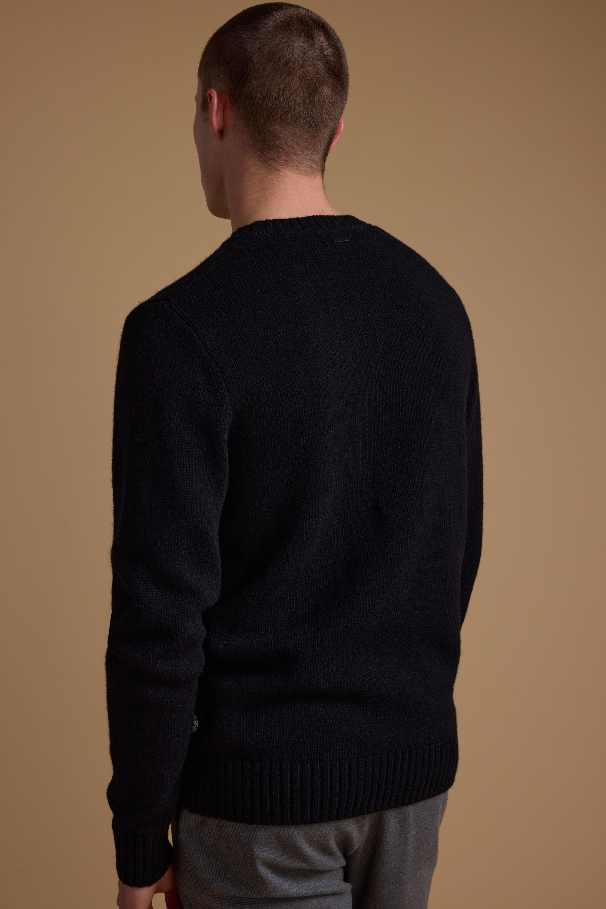 A person with short hair faces away from the camera, wearing Barkers' Anglers Merino Crew sweater in black and gray pants—a classic winter staple—set against a plain beige background.