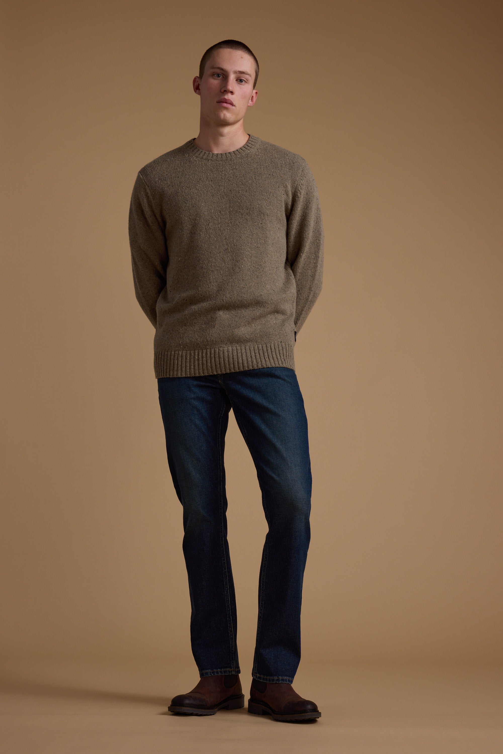 A person stands against a plain beige background, wearing the Barkers Anglers Merino Crew sweater in brown with dark blue jeans and brown shoes. Hands are behind their back as they face the camera with a neutral expression.