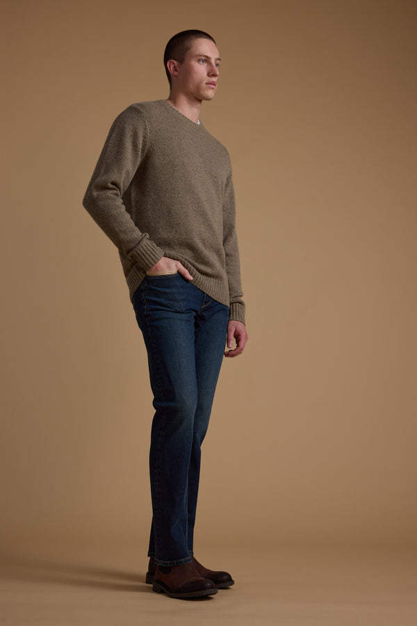 A man stands sideways with one hand in his pocket, wearing the Barkers Anglers Merino Crew sweater with blue jeans and brown boots against a plain tan background.