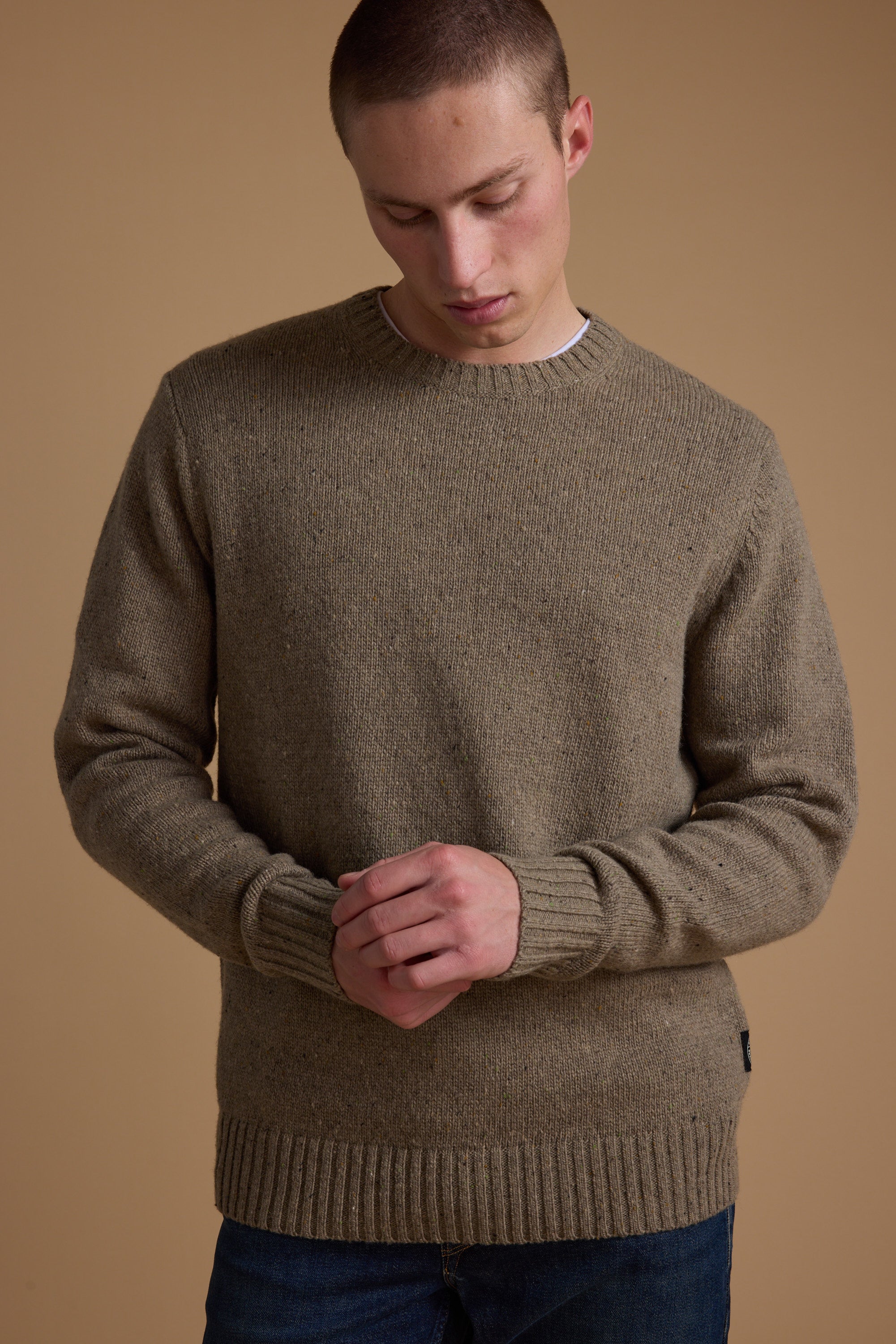 A young man with short hair wears the Barkers Anglers Merino Crew sweater and blue jeans, standing against a beige background with his hands clasped—a timeless winter staple.
