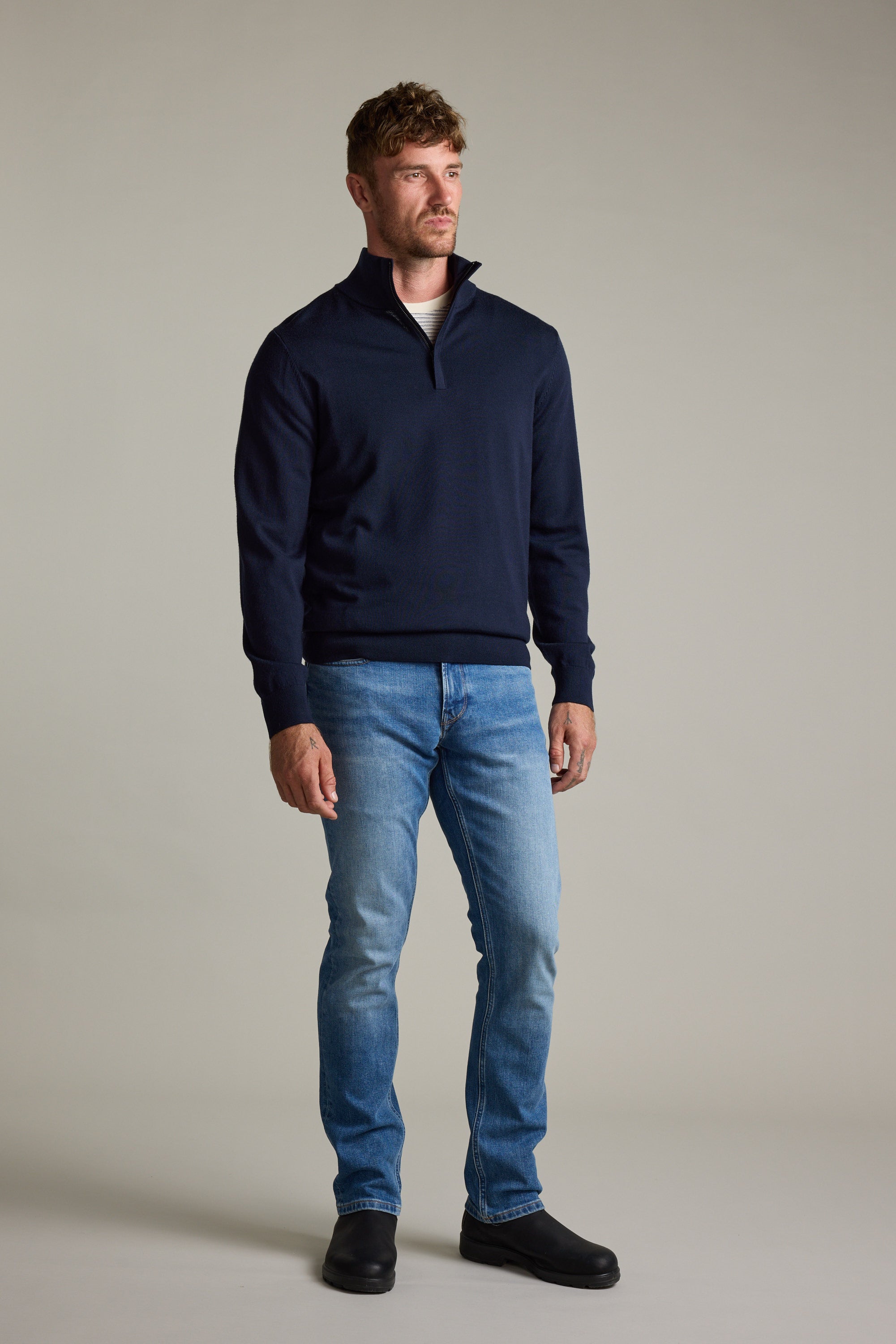 Man wears Barkers Skyland ZQRX Merino 1/4 Zip, blue jeans, and black shoes against plain background.