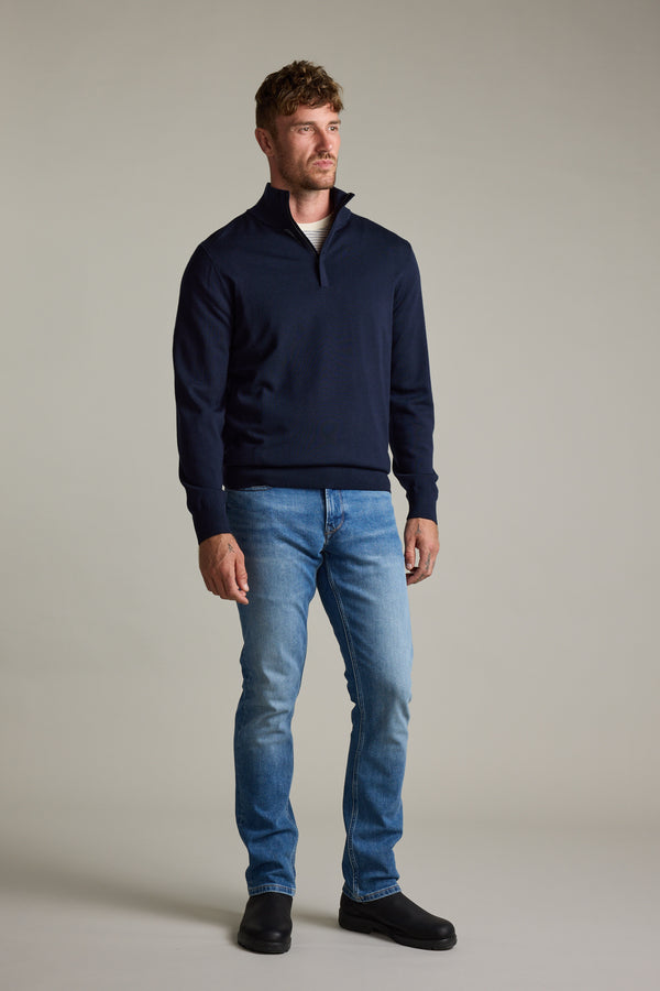 Man wears Barkers Skyland ZQRX Merino 1/4 Zip, blue jeans, and black shoes against plain background.